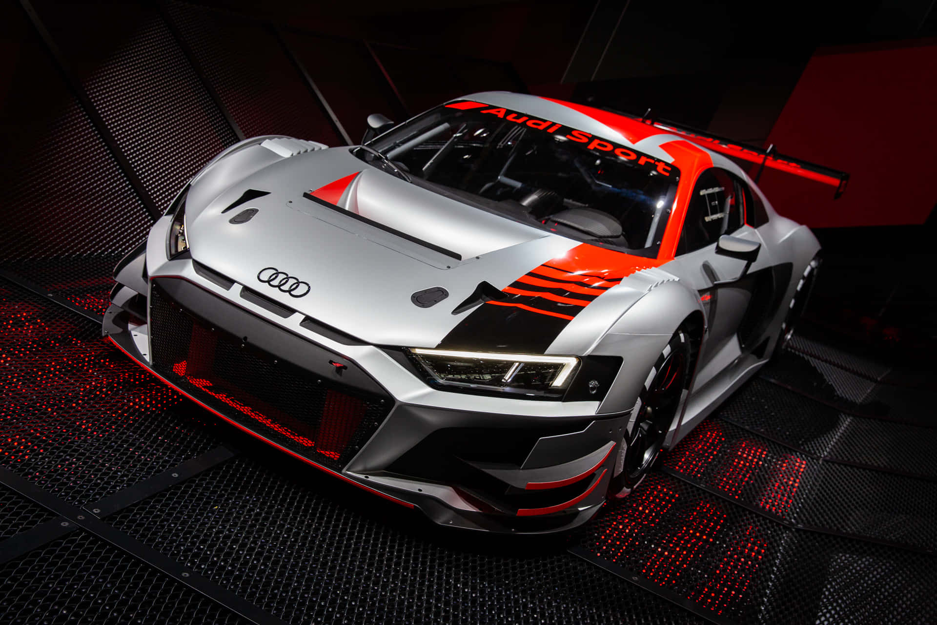 以奧迪提升您的駕駛體驗 壁紙 — 一架時尚的銀色奧迪 R8 LMS GT3 賽車，帶有鮮明的紅色點綴和發光的底盤燈，停在光線昏暗的車庫中的黑色穿孔地板上。