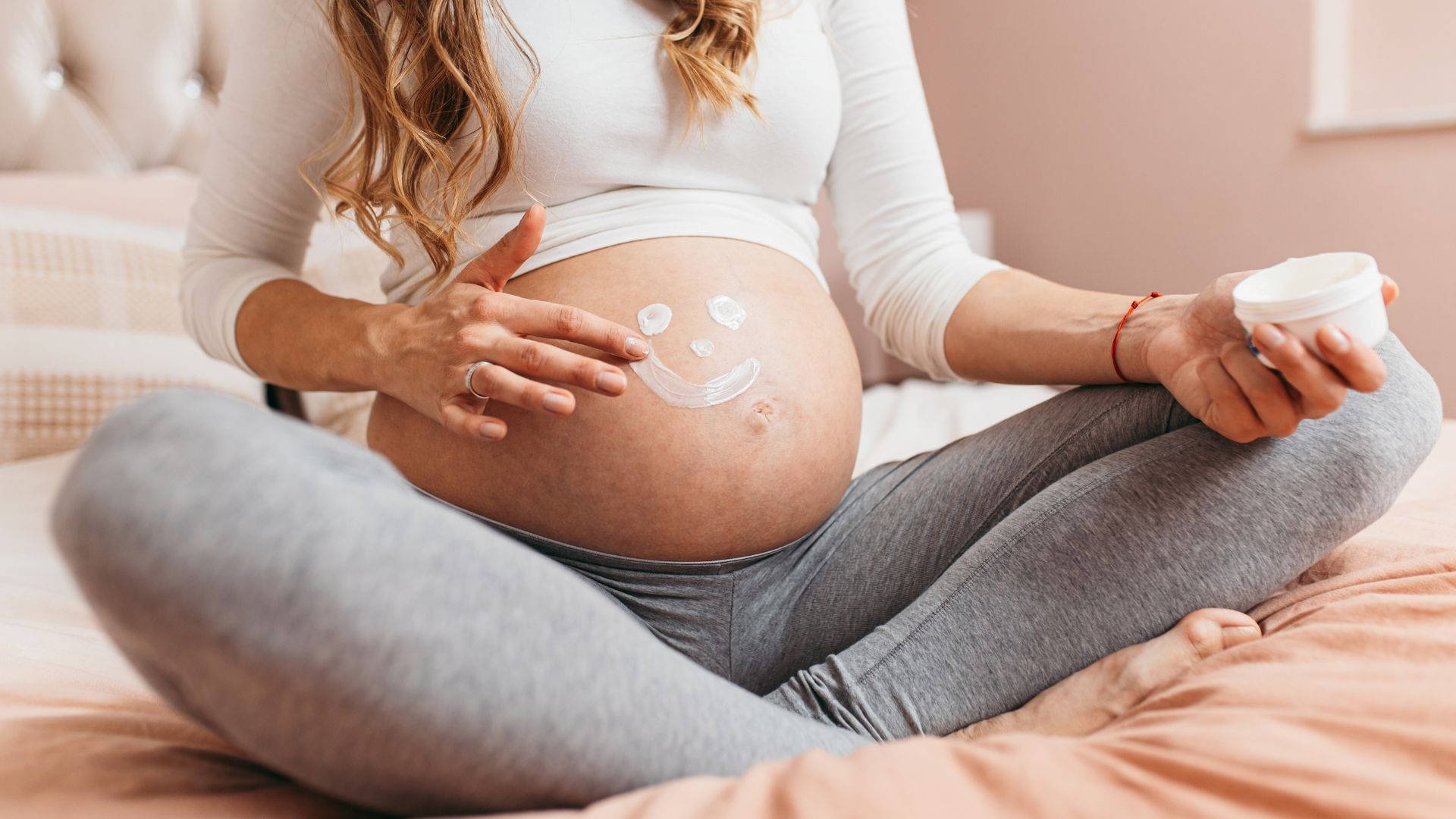 孕婦坐在床上，在肚子上塗抹乳液形成笑臉形狀。