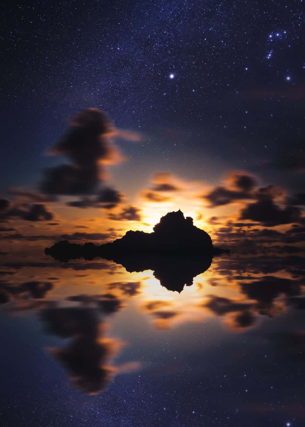 在繁星點點、雲朵發光的夜空下，岩石島嶼的輪廓倒映在水中。