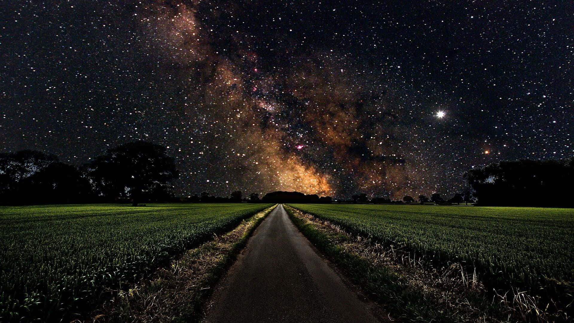 以完美的繁星夜空背景探索夜空之美 壁紙 — 一條黑暗蜿蜒的道路穿過綠色的田野，上方是令人驚嘆的星空，銀河在頭頂閃耀。