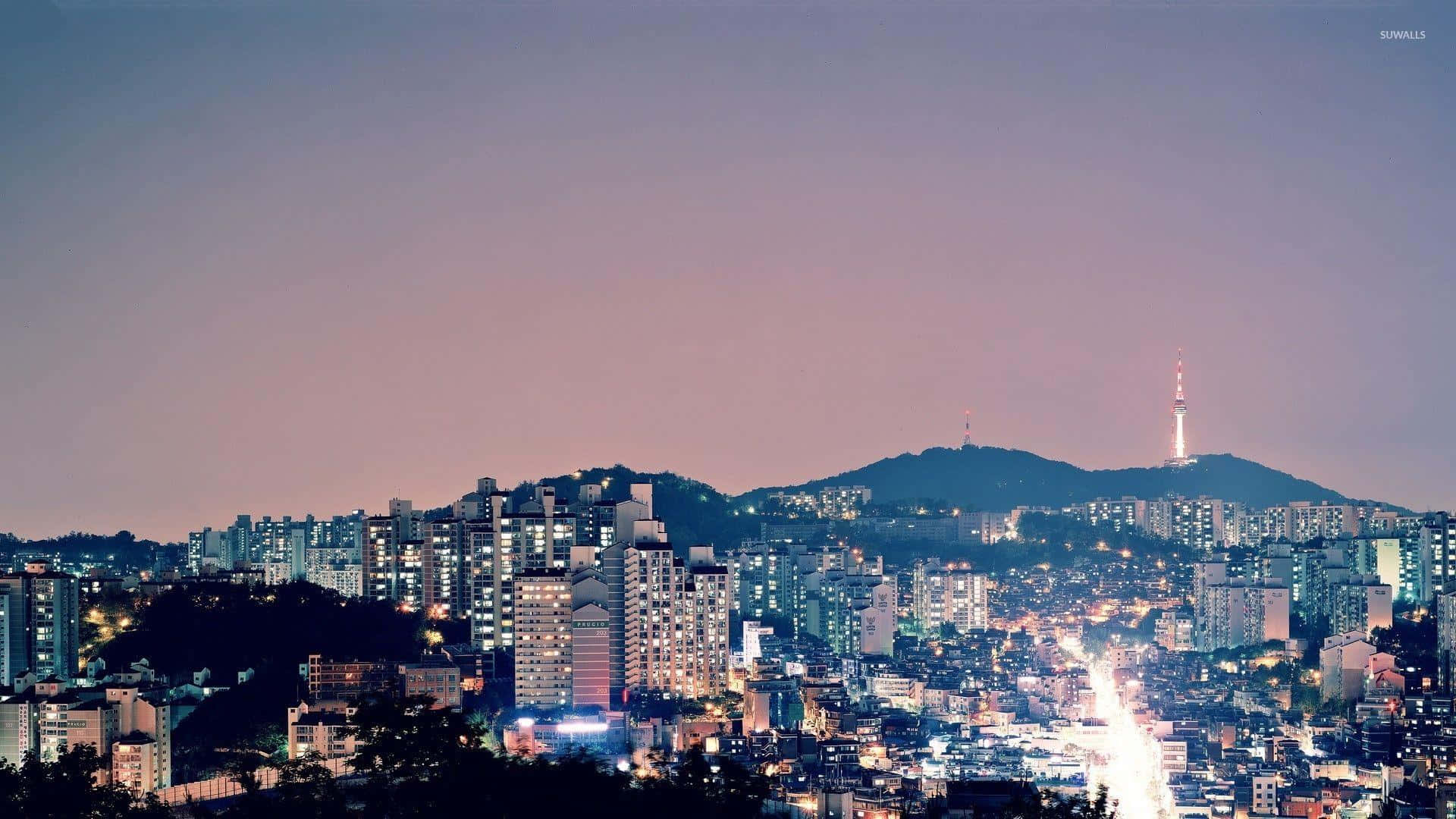 黃昏時分的城市天際線，建築物燈火通明，山丘上有一座塔樓。