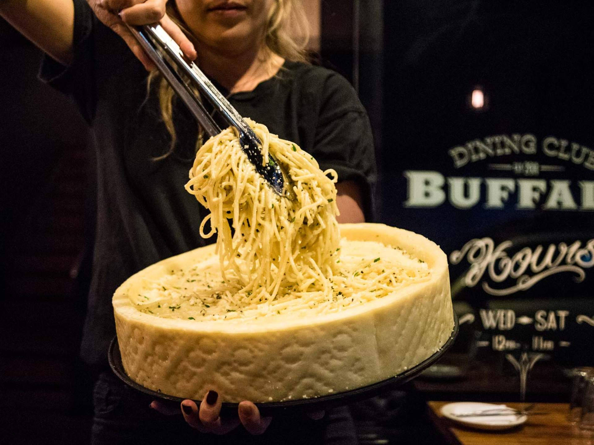 以帕瑪森起司輪盛裝的 Cacio E Pepe 壁紙 — 在燈光昏暗的餐廳裡，服務員用夾子從一個巨大的帕瑪森芝士輪中提起意大利麵。