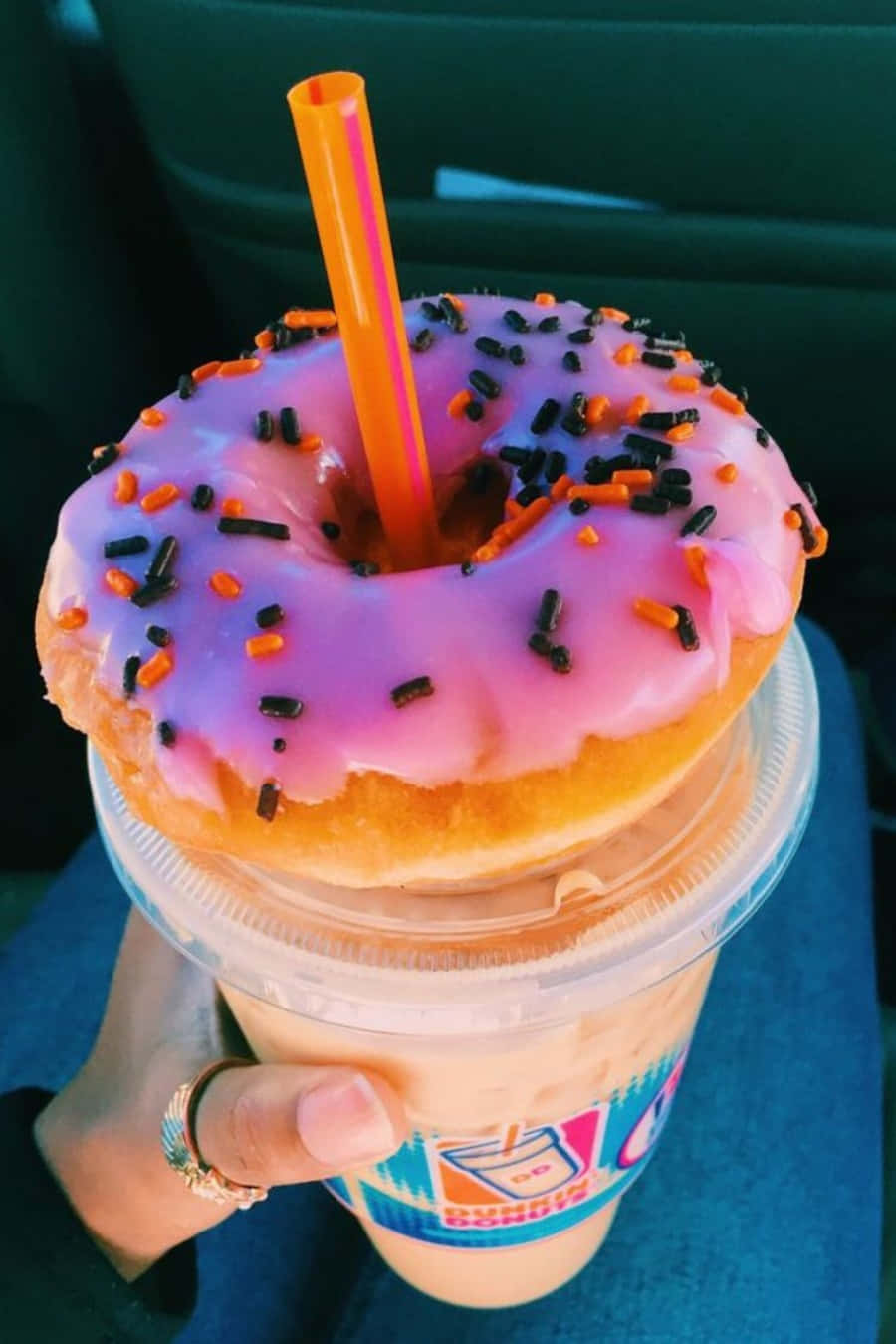 以微笑開始一天，選擇Dunkin' Donuts 壁紙 — 一隻手拿著一杯奶油冰飲，上面放著一個粉紅色糖霜甜甜圈，撒有黑色和橙色的糖針。