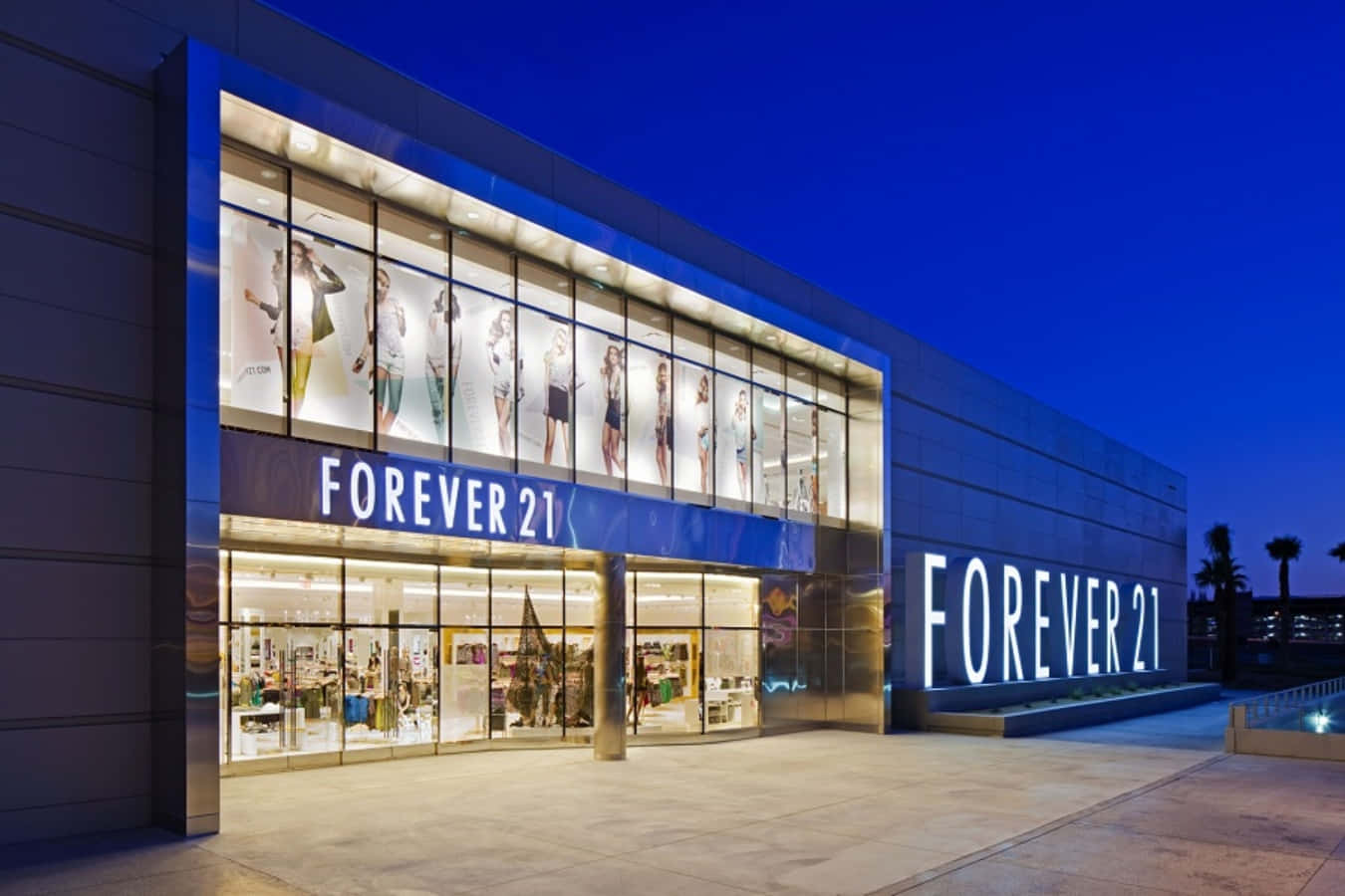 以時尚又實惠的Forever 21更新衣櫥 壁紙 — 暮色中燈火通明的Forever 21商店外牆，明亮的櫥窗展示著人體模型和醒目的招牌，襯托著深藍色的天空。