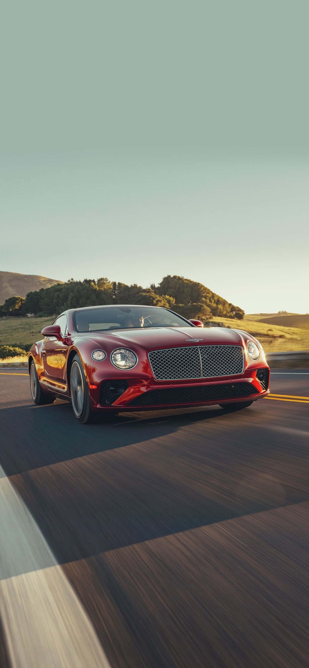 以時尚新 iPhone XS Max 與 Bentley 體驗奢華 壁紙 — 一架流線型紅色賓利 Continental GT 在晴朗天空下沿著蜿蜒道路穿過金色山丘飛馳。