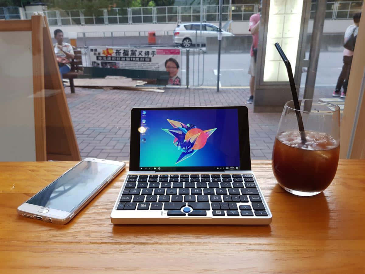 以時尚現代筆電提升生活品質 壁紙 — 一部帶有彩色摺紙鳥壁紙的緊湊型筆記本電腦放在咖啡廳的木桌上，旁邊是一部智能手機和冰咖啡。