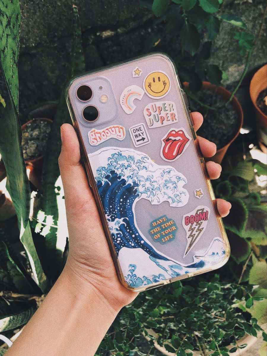 以時尚美學 iPhone 升級您的造型 壁紙 — 一隻手拿著一個裝飾有彩色貼紙和藍色波浪設計的手機殼，背景是茂盛的綠色盆栽植物。