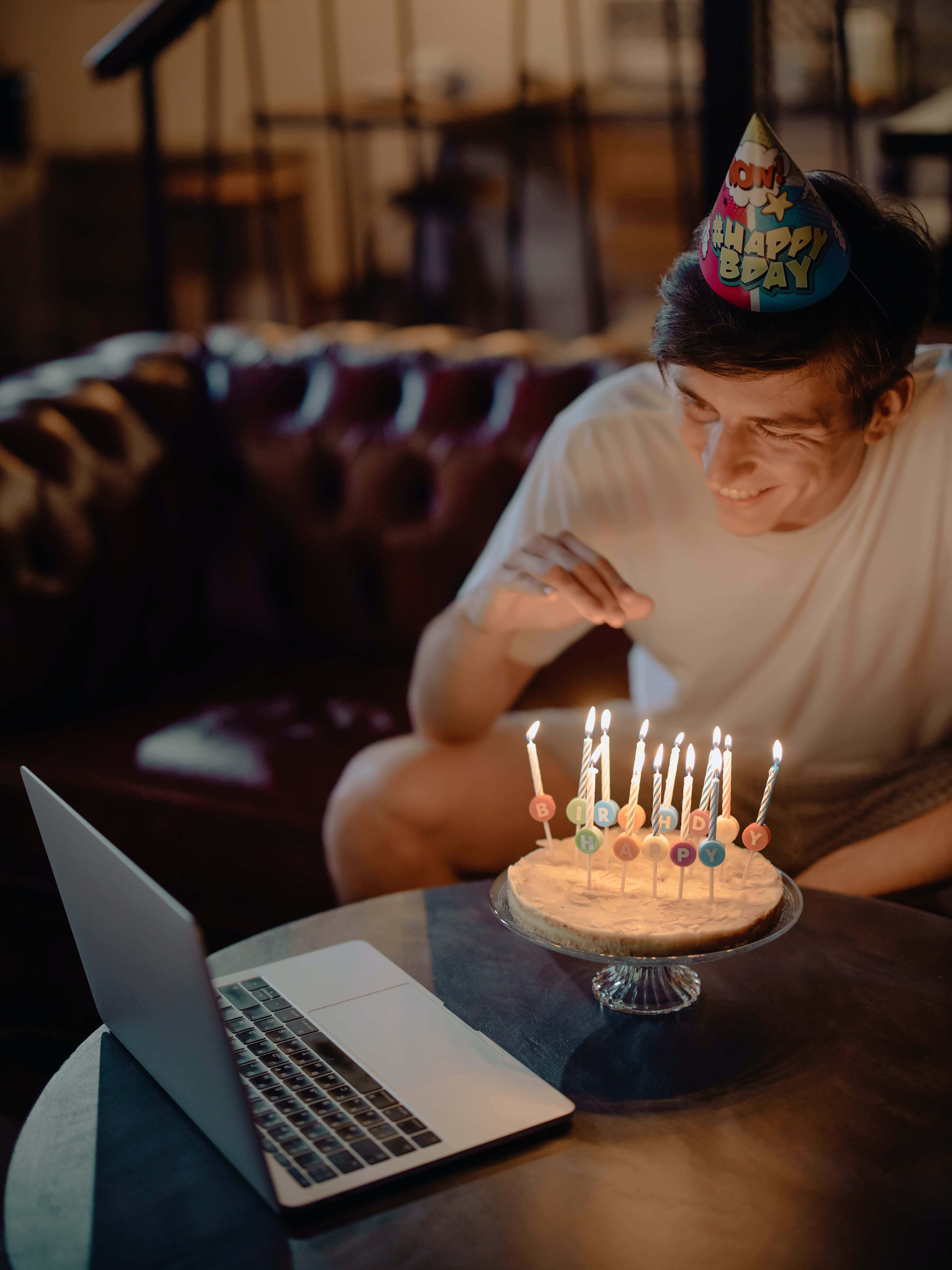 以無憂無慮的喜悅慶祝你的特別日子 壁紙 — 一位戴著生日帽的年輕男子，一邊在筆記型電腦上進行視訊通話，一邊微笑著吹熄蛋糕上的蠟燭。