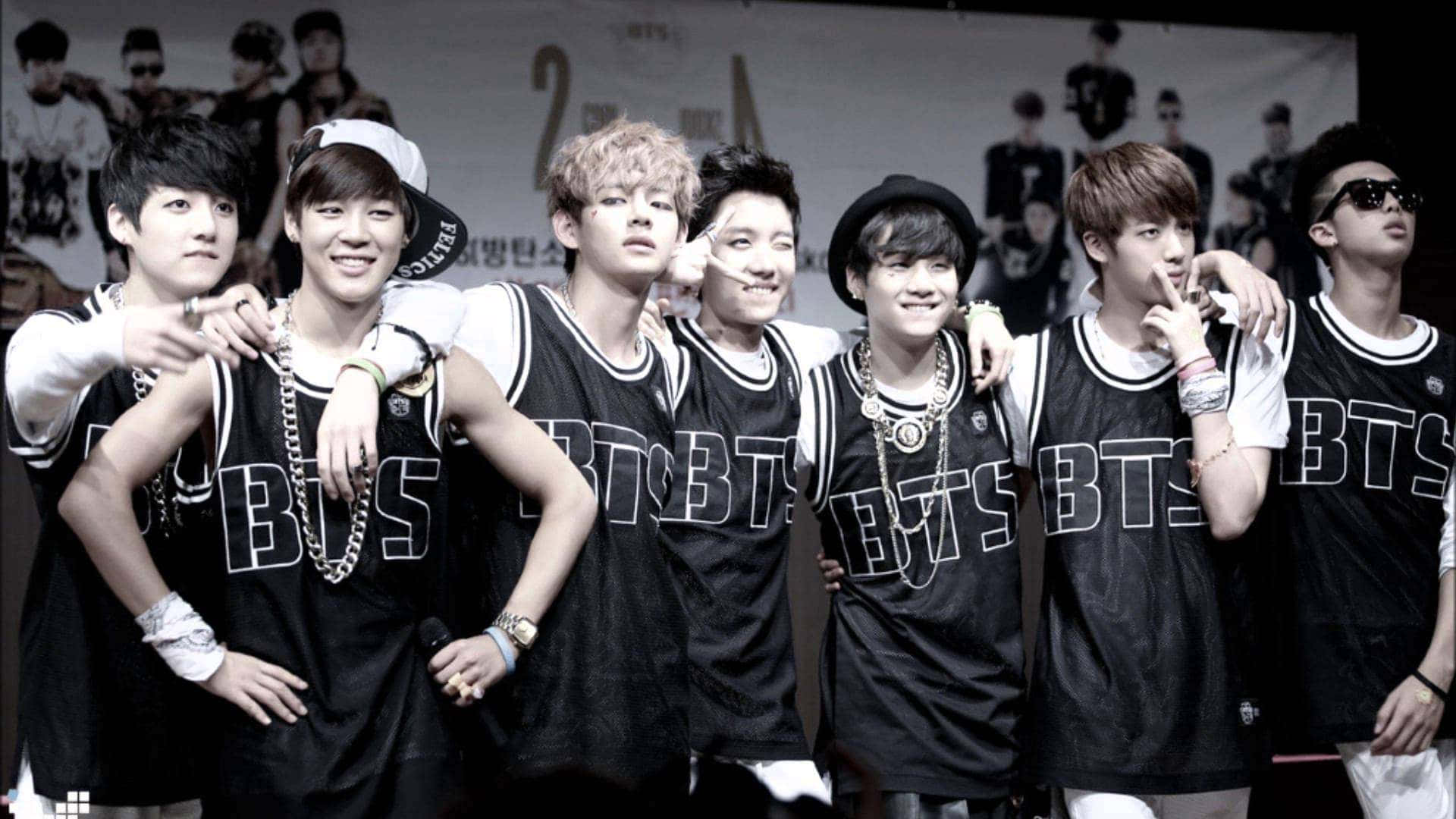 以熱情與熱忱慶祝韓流文化 壁紙 — 七位身穿黑色「BTS」球衣的年輕男子，戴著鏈條和帽子，在宣傳背景前自信地擺姿勢。