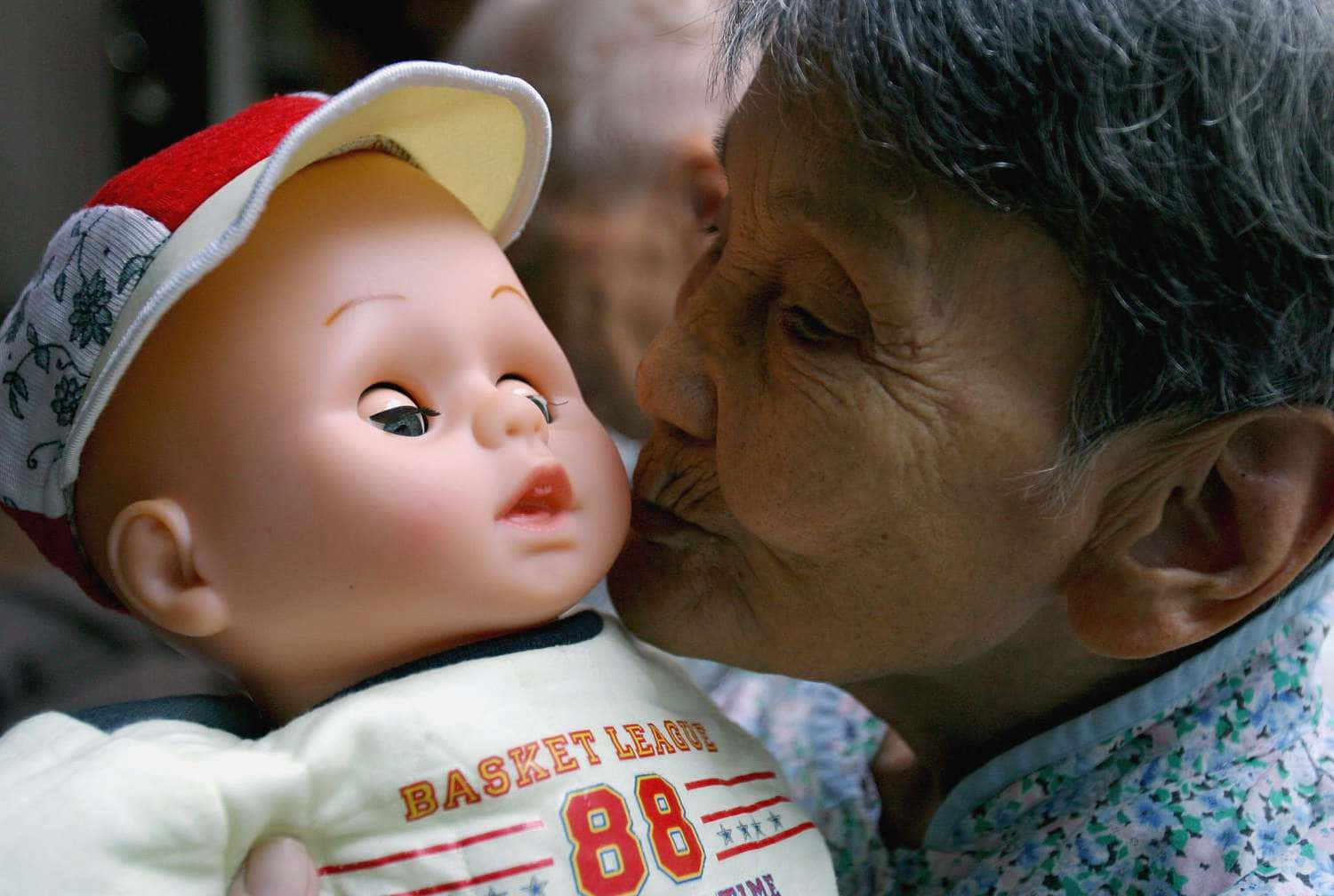 以玩偶慶祝愛與奉獻 壁紙 — 一位年長女性溫柔地親吻一個戴著紅白帽子和穿著「Basket League 88」襯衫的嬰兒娃娃。