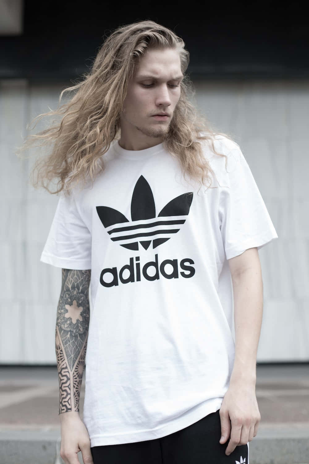 以現代詮釋經典 Adidas 風格 壁紙 — 一位年輕男子，長著波浪長髮和紋身手臂，穿著白色Adidas T恤，背景是樸素的城市風光。