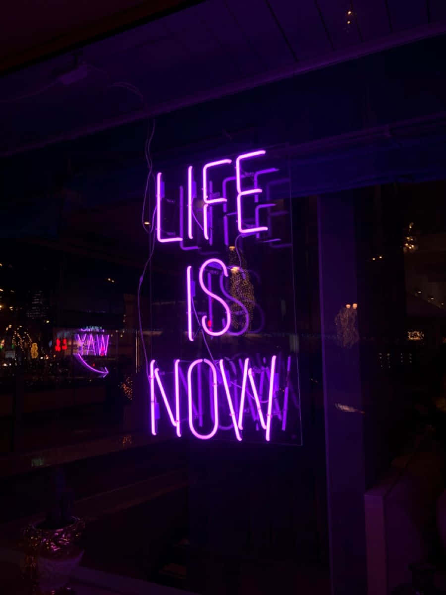夜晚黑暗的城市窗戶中，一個寫著「Life is Now」的紫色霓虹燈牌。
