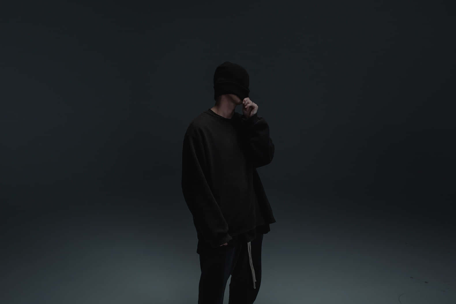 一個穿黑色衣服的人站在黑暗的攝影棚中，手靠近臉部。