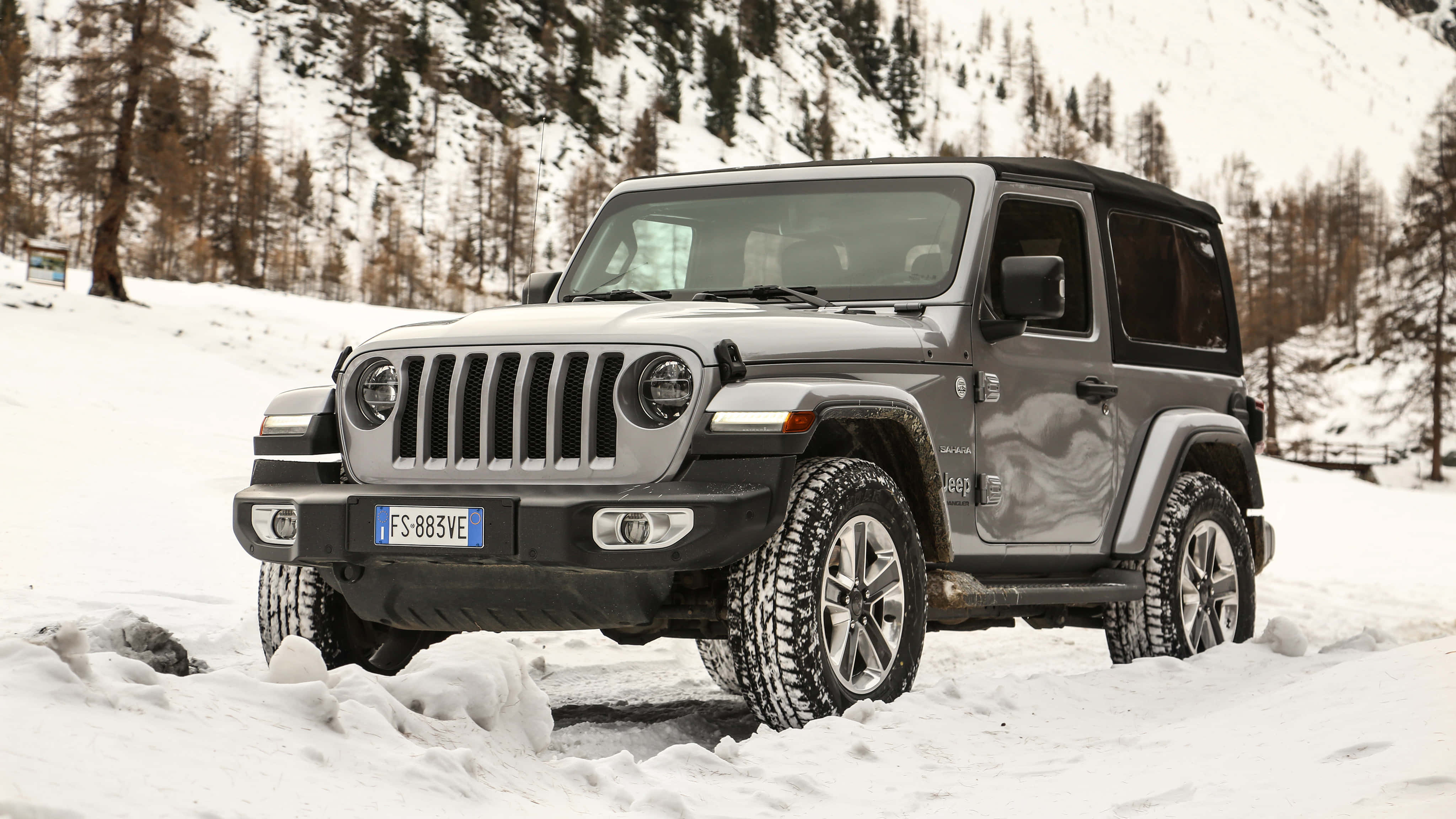 以經典Jeep Wrangler啟動你的下一次冒險 壁紙 — 一輛灰色Jeep Wrangler在積雪深厚的雪地中行駛，附近有被雪覆蓋的松樹和岩石山腰。