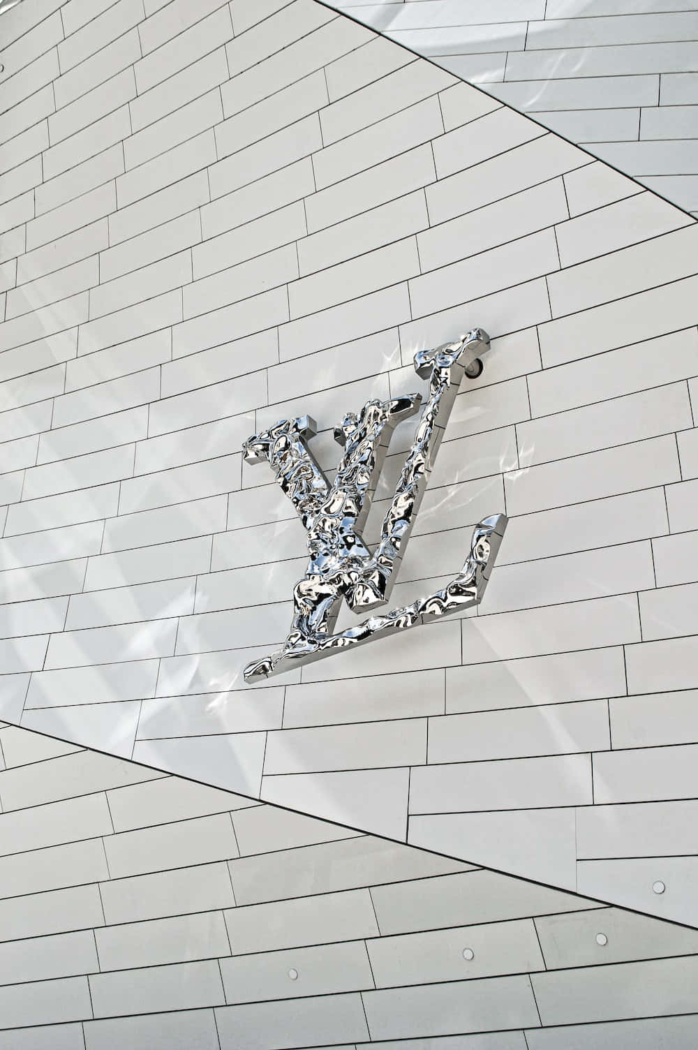 以經典路易威登展現獨特風采 壁紙 — 一個時尚的鉻色 Louis Vuitton 標誌安裝在白色瓷磚牆上，帶有幾何角度和銳利陰影。