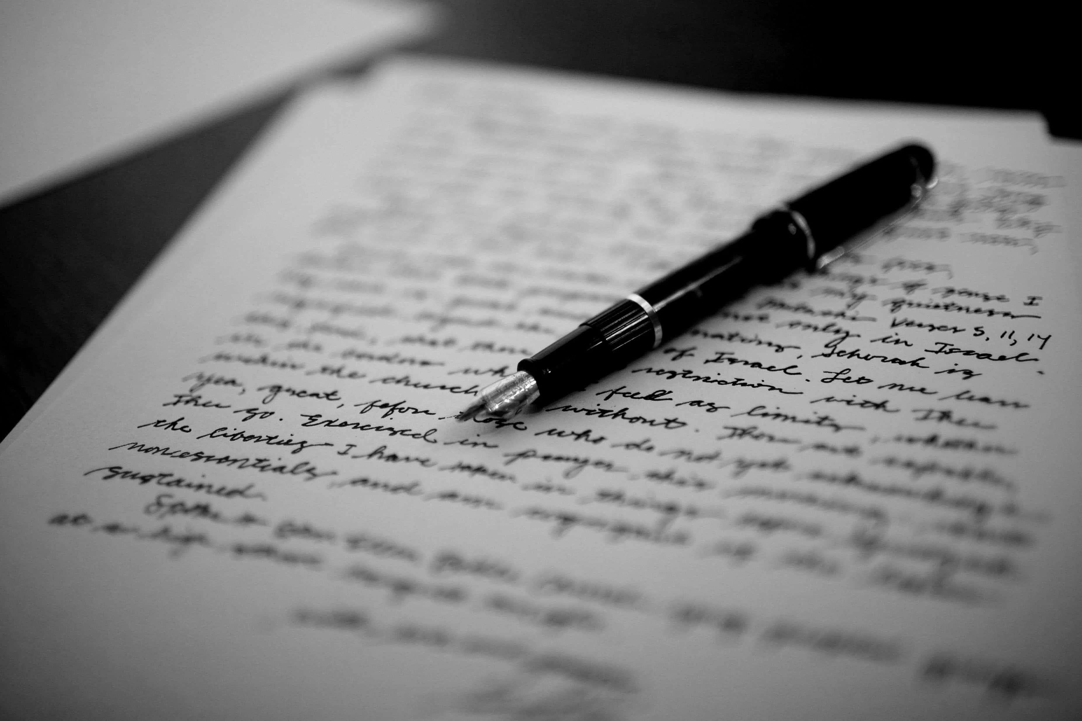 以經典鋼筆為美好時刻點綴 壁紙 — 一支黑色鋼筆放在手寫字頁面上，呈現淺景深黑白效果。