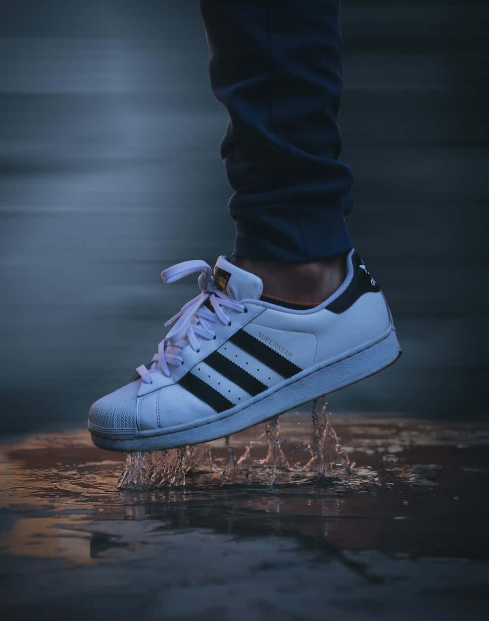 以經典阿迪達斯鞋履提升你的表現 壁紙 — 一對白色 adidas superstar 運動鞋在淺水中濺起水花，水滴在空中凝固，背景模糊黑暗。