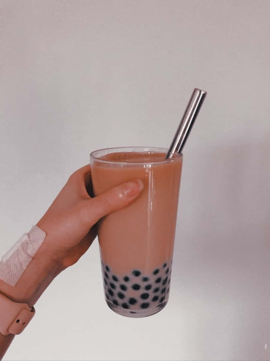 以美味珍珠奶茶解渴 壁紙 — 一隻手拿著一杯高高的奶油粉紅泡泡茶，裡面有黑色木薯珍珠和金屬吸管，背景是純白色。