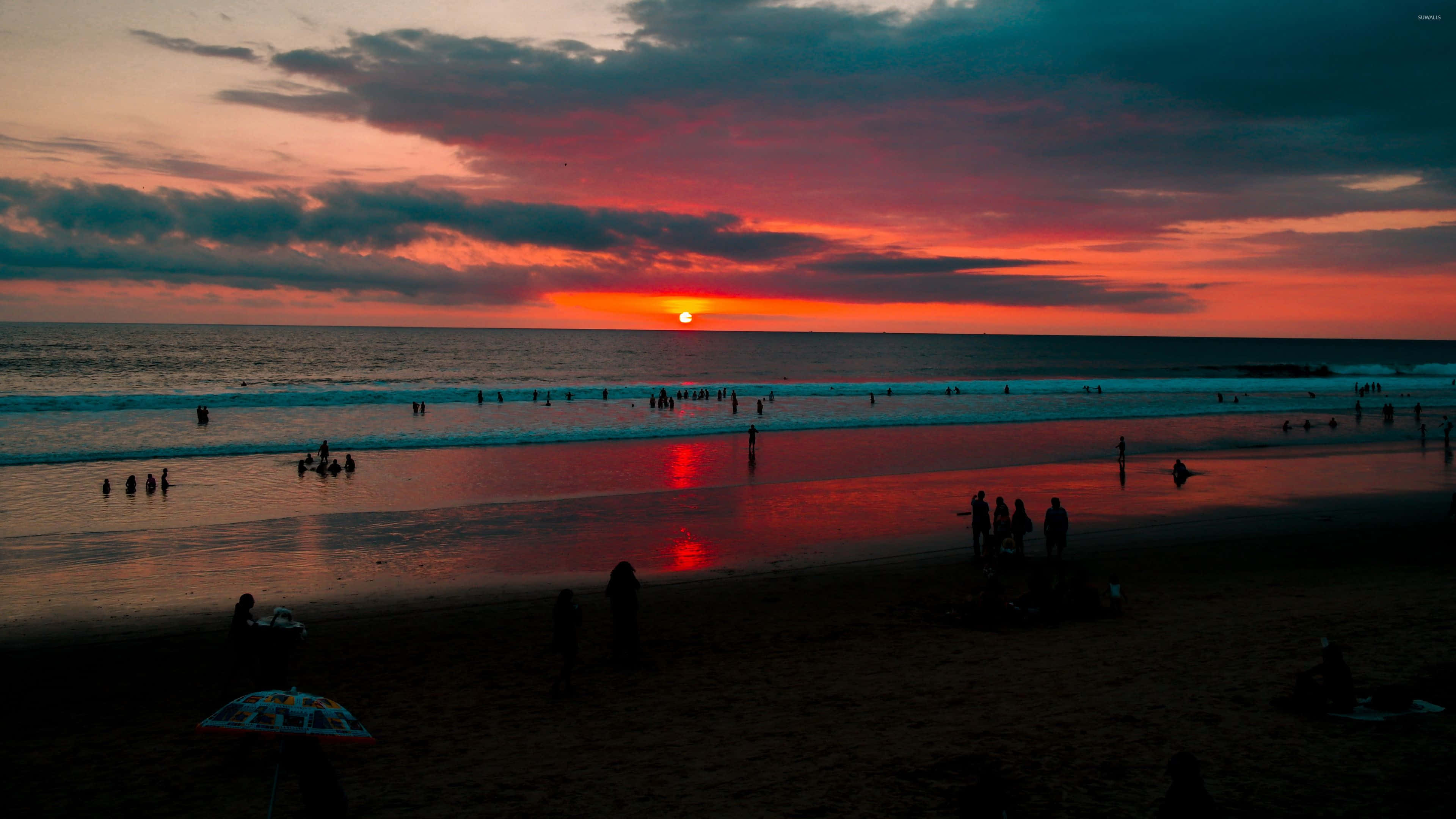 以美麗的海灘日落開始一天 壁紙 — 夕陽將天空染成火紅和橙色，海灘上的人們在剪影中享受海浪。