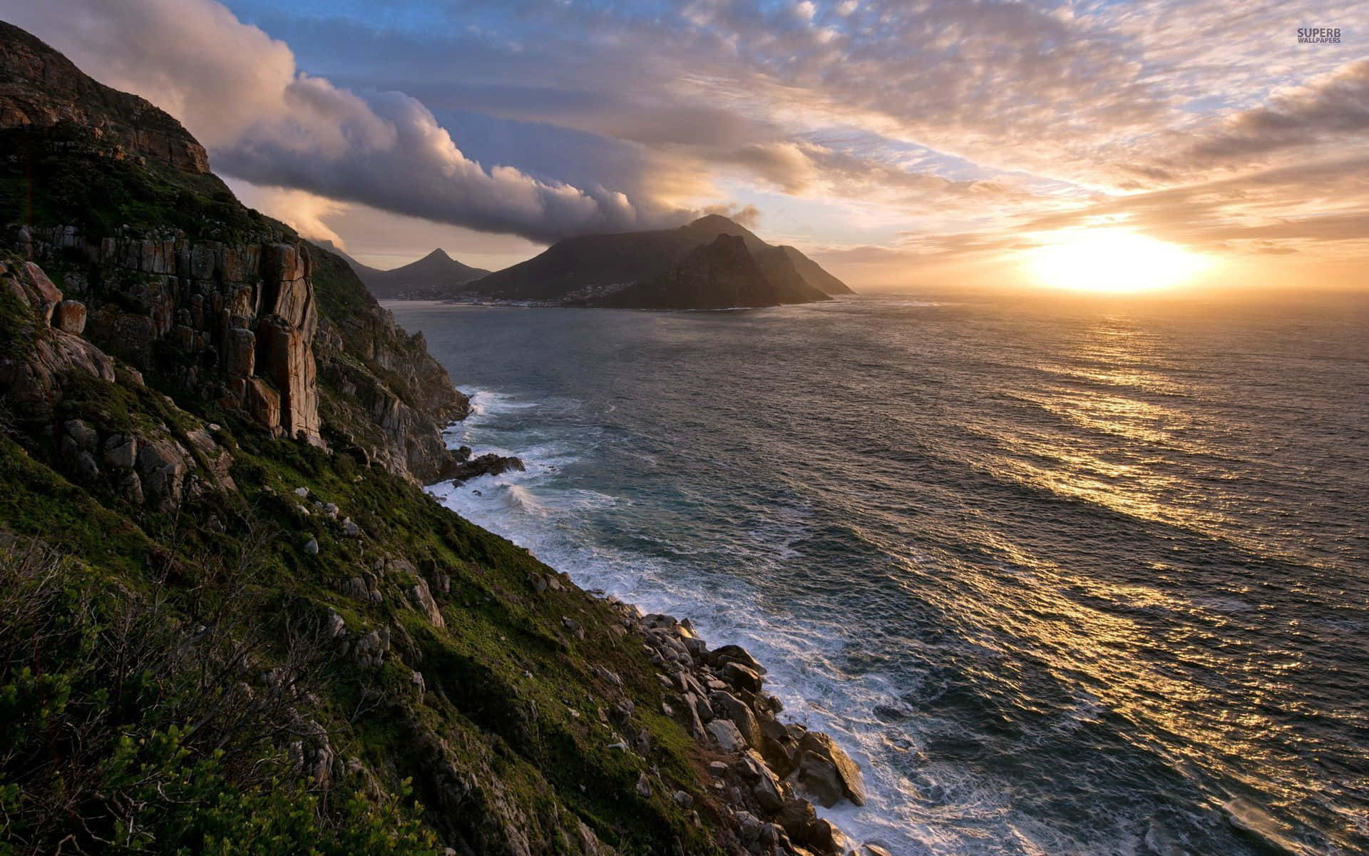 以美麗陽光開啟美好一天 壁紙 — 夕陽將海洋染成金色，海浪在戲劇性的雲層下拍打崎嶇的懸崖。