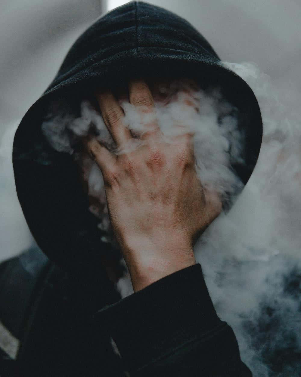 穿著黑色連帽衫的人用手遮住臉，白色蒸氣在周圍翻騰。