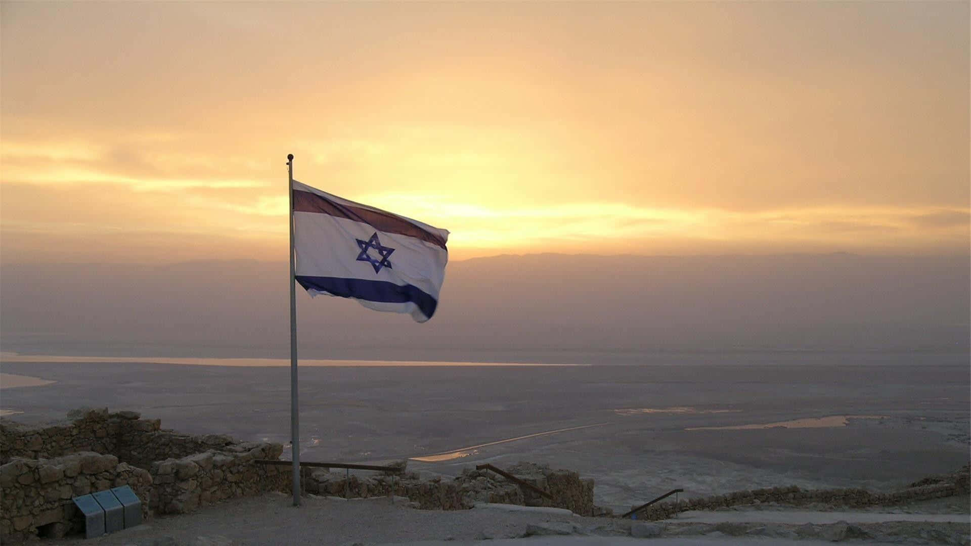 Israel 桌布 壁紙 — 以色列國旗在死海廣闊鹽灘上金色日落時分，自豪地飄揚在石頭觀景台上。