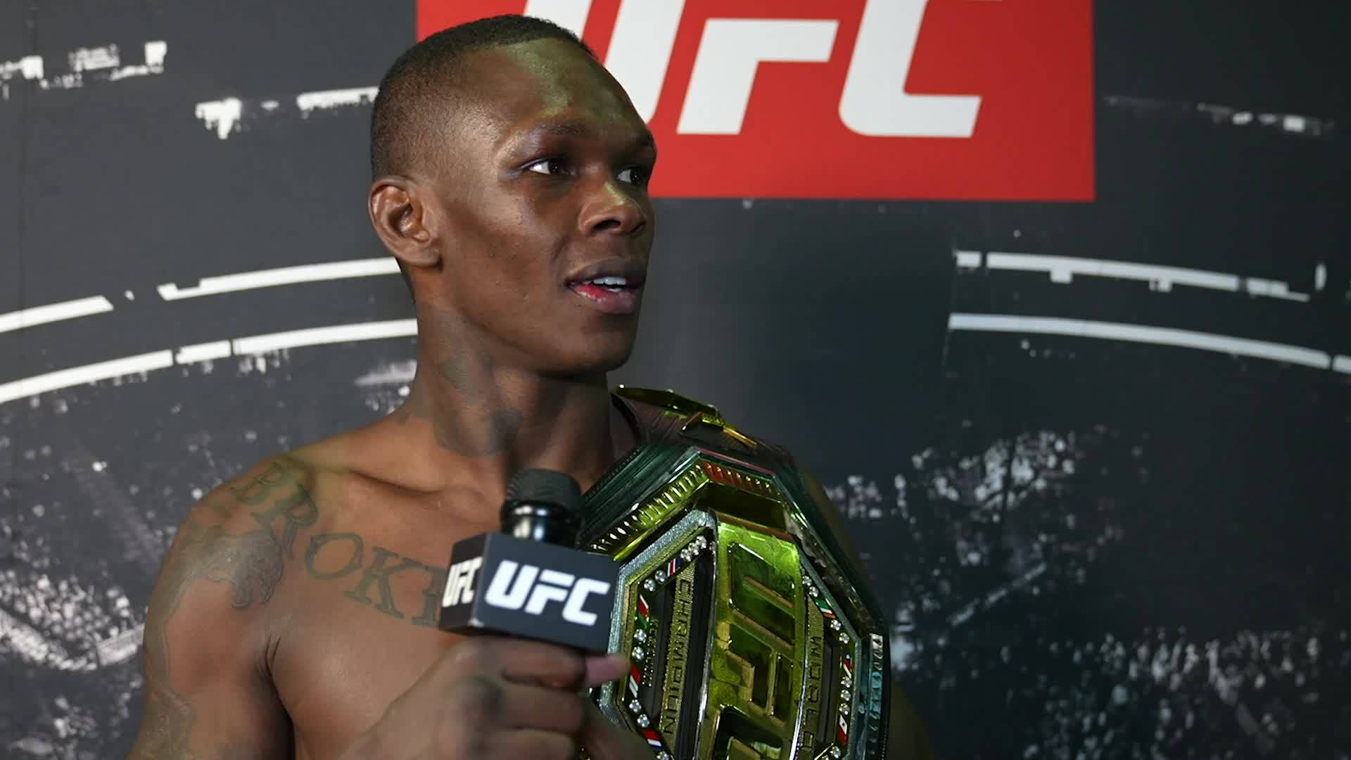 以色列阿德esan亞 UFC訪問 壁紙 — 一位肌肉發達的UFC拳手在記者會上拿著冠軍腰帶對著麥克風講話。