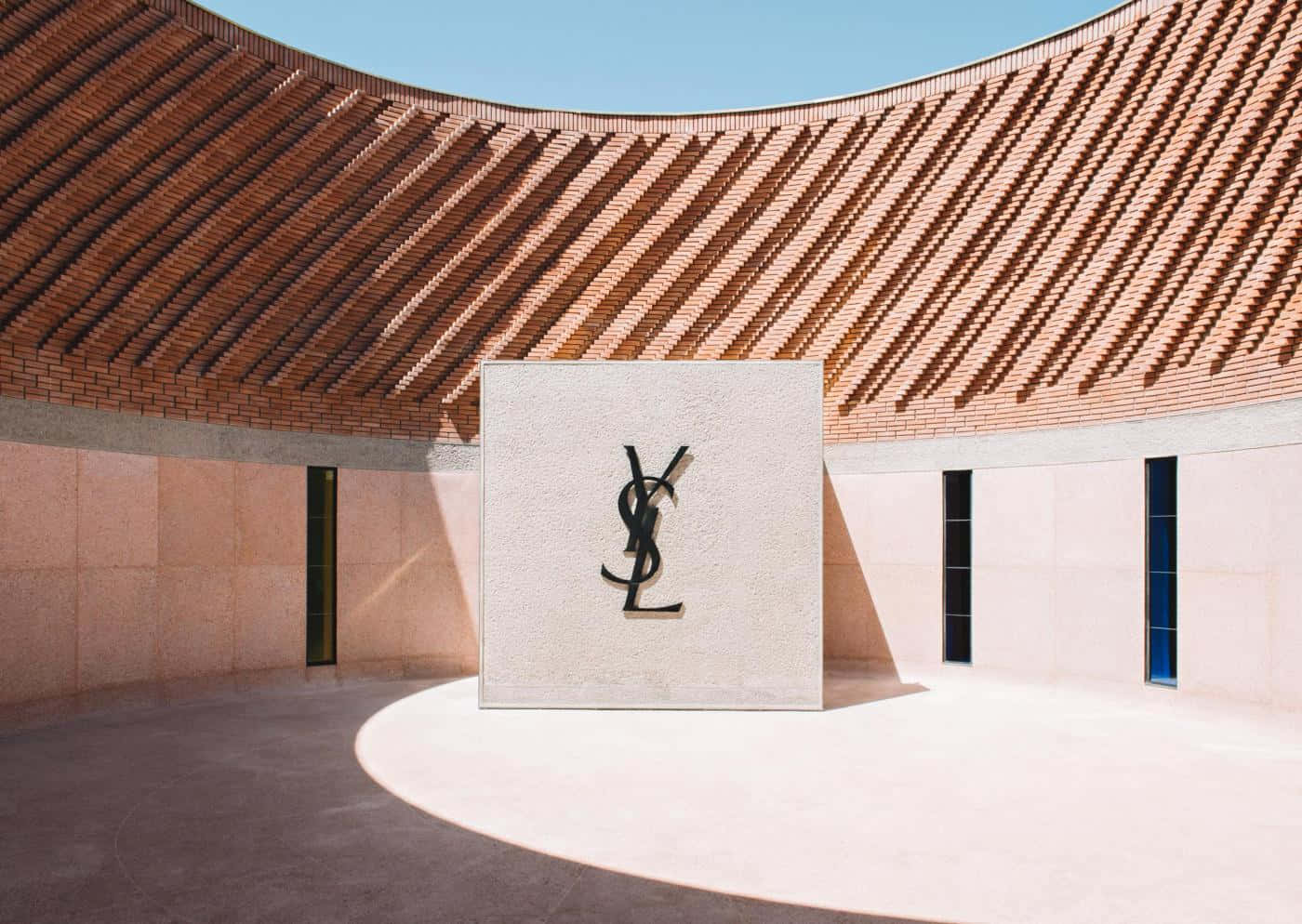 「以色彩活出自我，YSL」 壁紙 — 一座簡約現代建築，擁有弧形陶土屋頂，在陽光普照的庭院中投射出幾何陰影，庭院中有一個突出的YSL標誌。