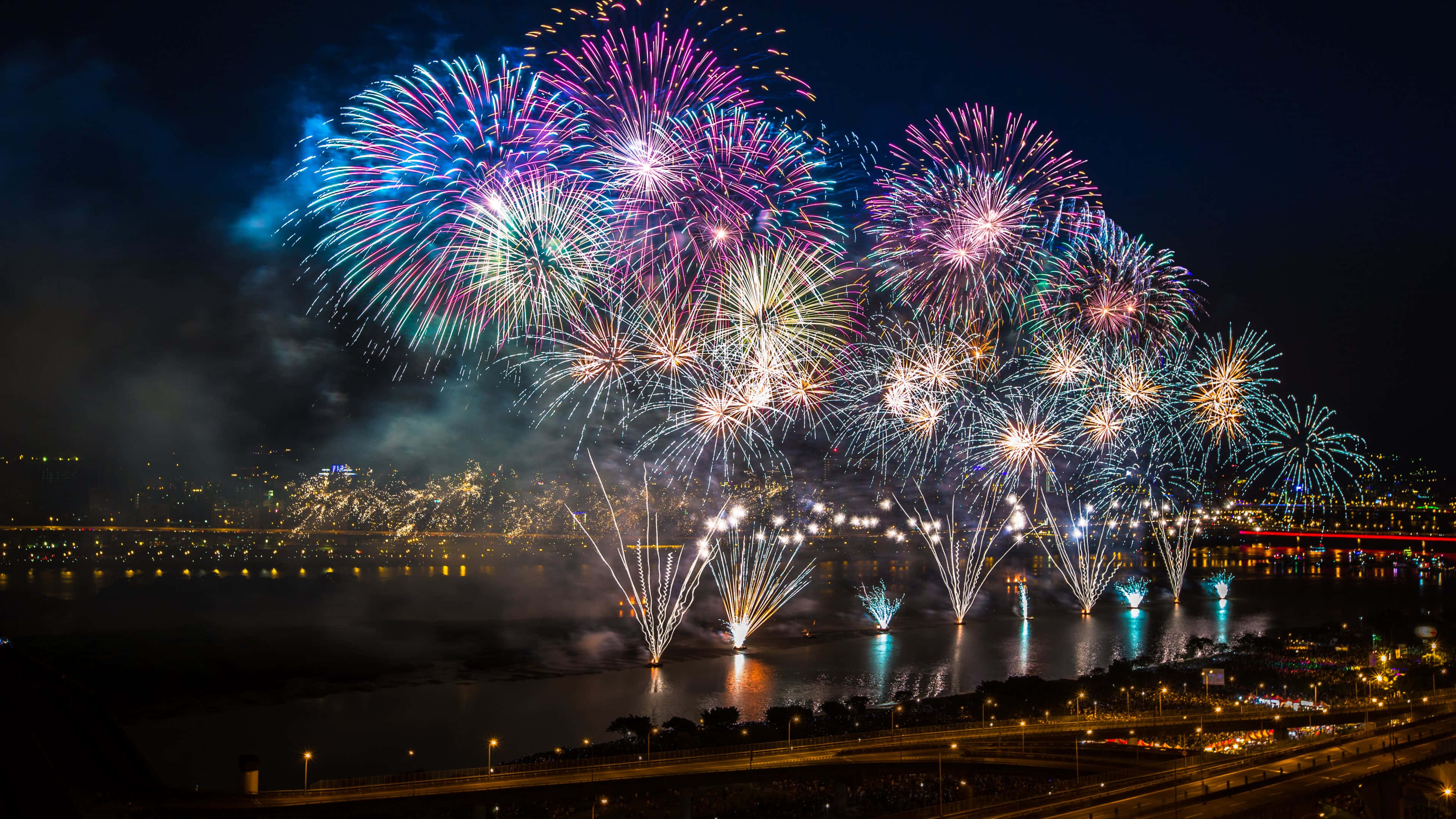 以色彩爆發慶祝！ 壁紙 — 充滿活力的煙火在夜間城市天際線倒映在黑暗水面上方綻放出耀眼色彩。