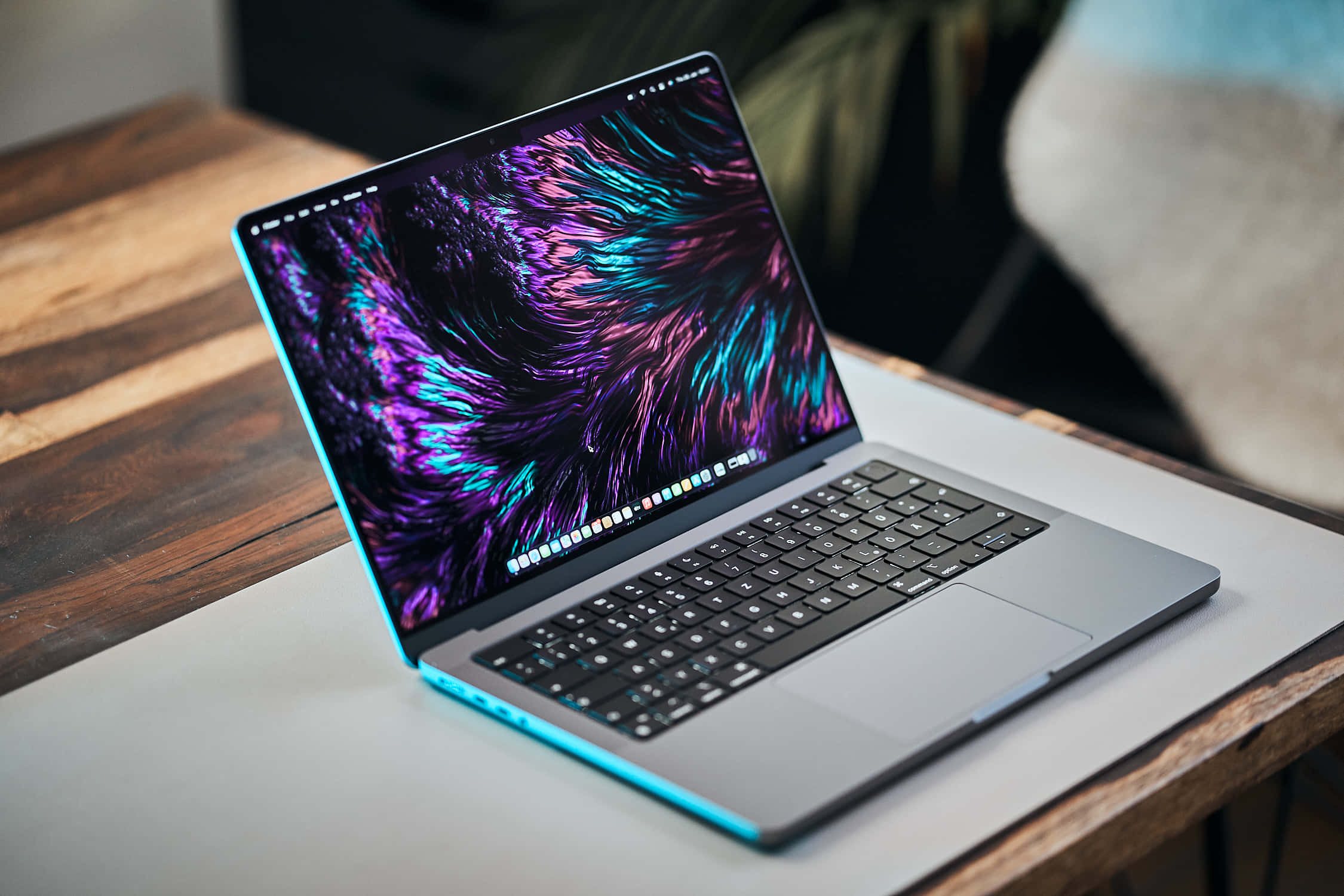 以藍色 MacBook 開啟您的筆記型電腦冒險 壁紙 — 一部時尚的銀色手提電腦，帶有發光的藍色邊緣，在木桌上顯示充滿活力的紫色和藍綠色抽象壁紙。