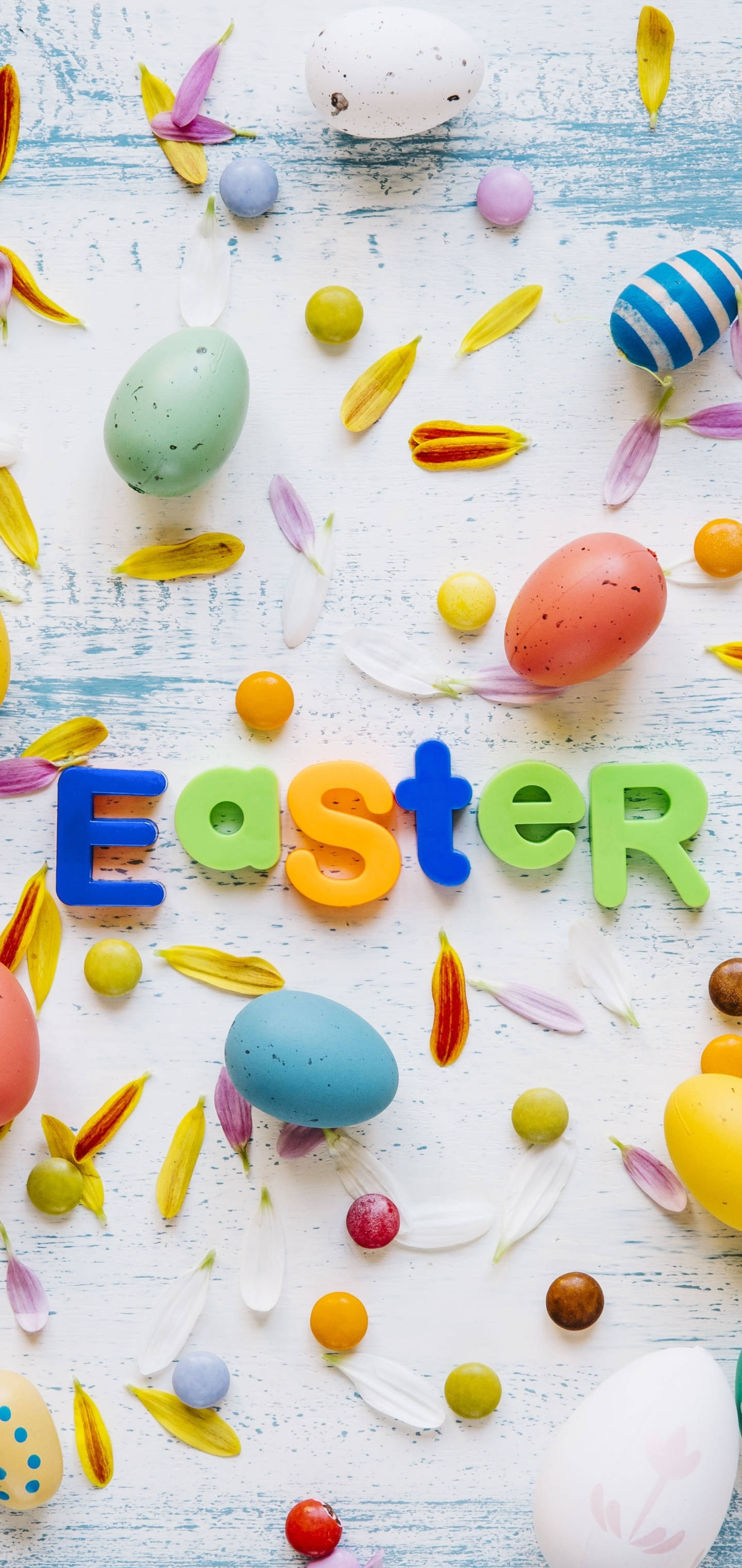 以詼諧微笑慶祝復活節！ 壁紙 — 色彩繽紛的復活節蛋和花瓣散落在風化的藍色木質表面上，並用明亮的字母拼出Easter一詞。