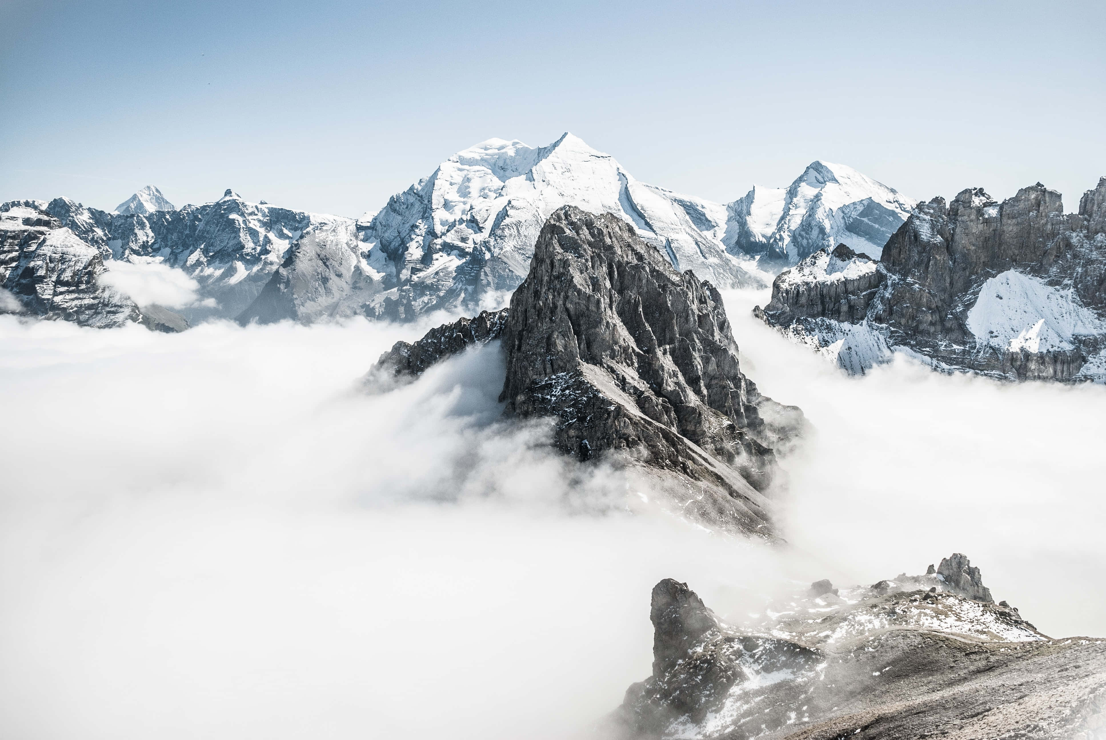 以這款免費背景攀上新高峰 壁紙 — 一座鋸齒狀的雪山峰在清澈的藍天下刺穿厚厚的雲層。