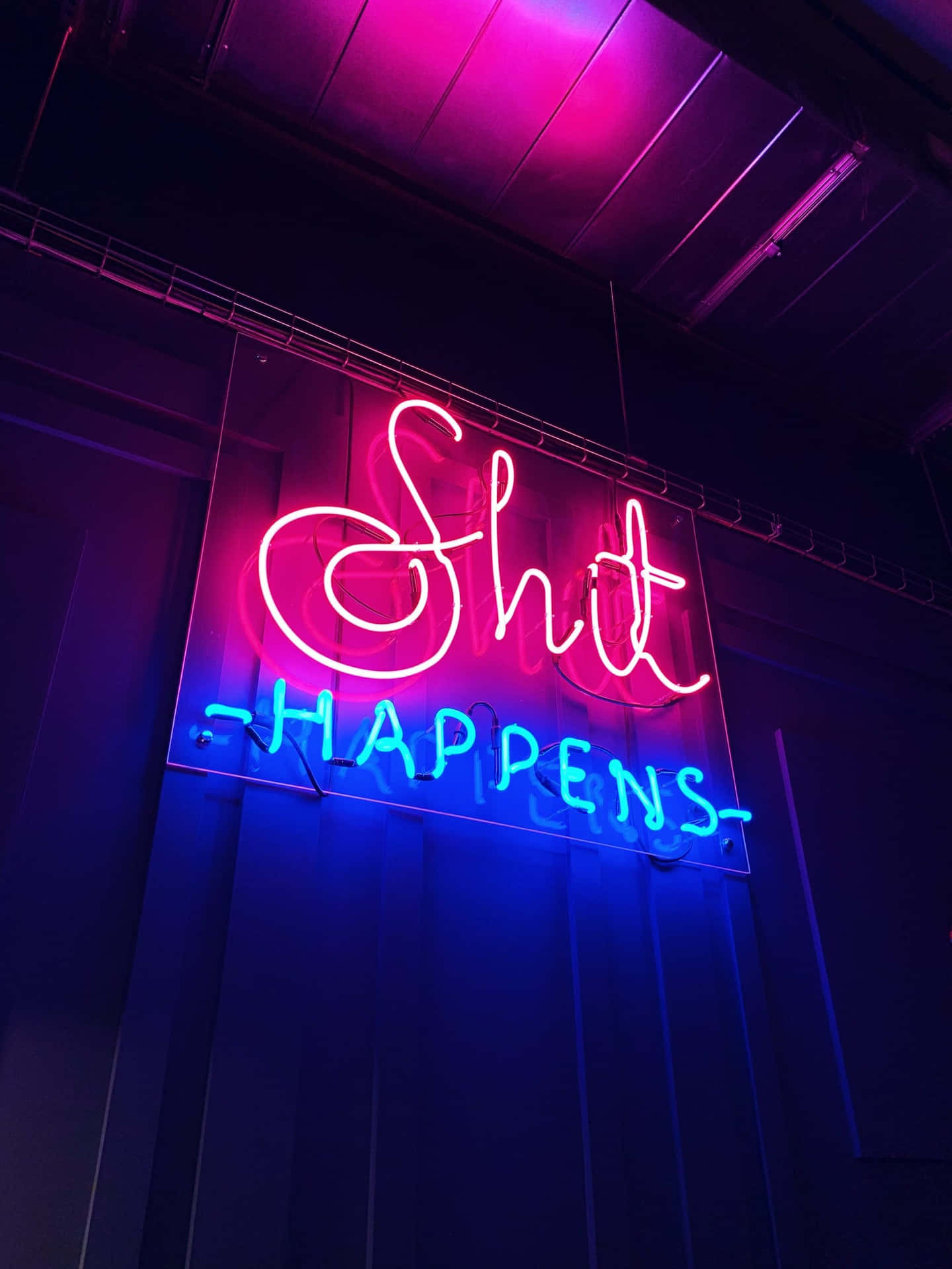 在黑暗的房間中，帶有粉紅色和藍色文字的霓虹燈牌，寫著 'Shit Happens'。