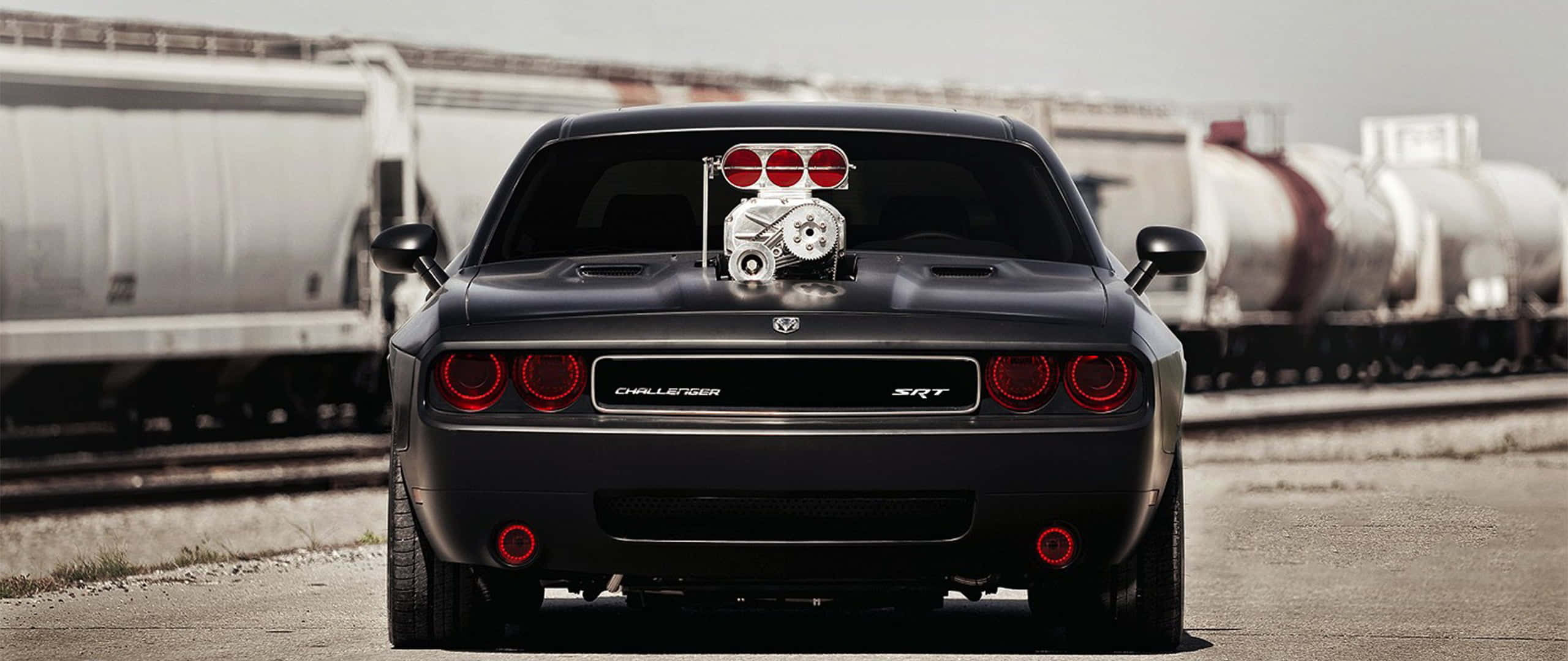 以道奇Hellcat主宰道路 壁紙 — 一輛黑色Dodge Challenger SRT，引擎蓋上裝有增壓器，停泊在火車軌道旁。