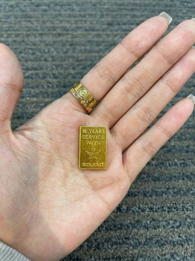 以金條穩固財富 壁紙 — 一隻手拿著一個刻有「10 YEARS SERVICE WITH saudia」的小金條，背景為帶紋理的灰色。