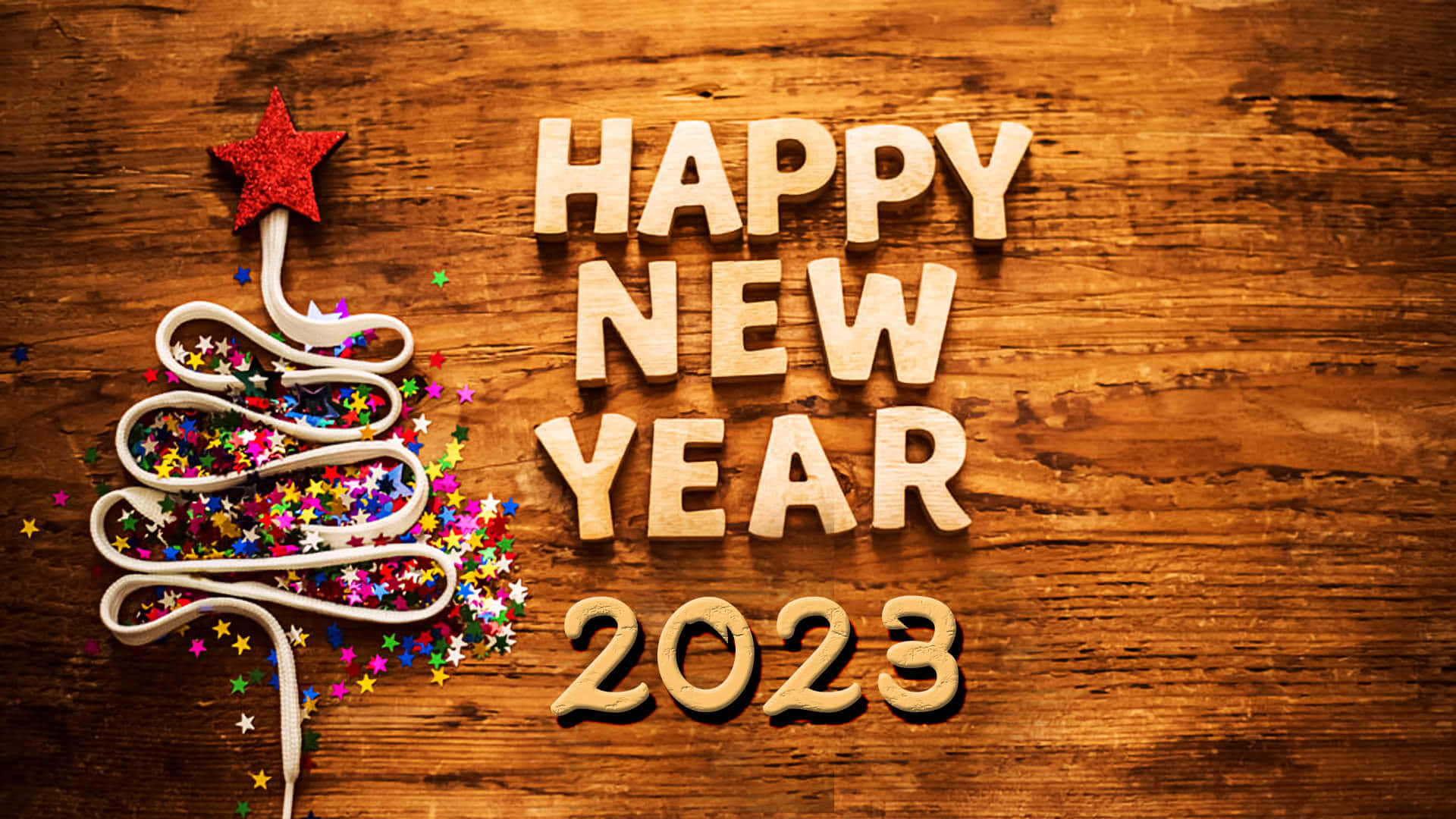 以開懷姿態迎接新年 壁紙 — 木製字母拼出新年快樂2023，旁邊有一個用絲帶捲成的樹，頂部有紅色星星，並在質樸的木頭上散落著閃粉。