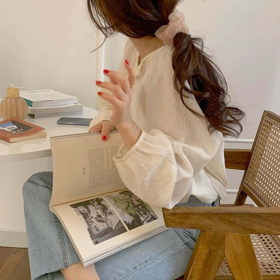 以雅緻米色融合自然與藝術之美 壁紙 — 一位年輕女性，紅色指甲，粉紅色髮圈，坐在白色書桌前的木椅上閱讀一本書。