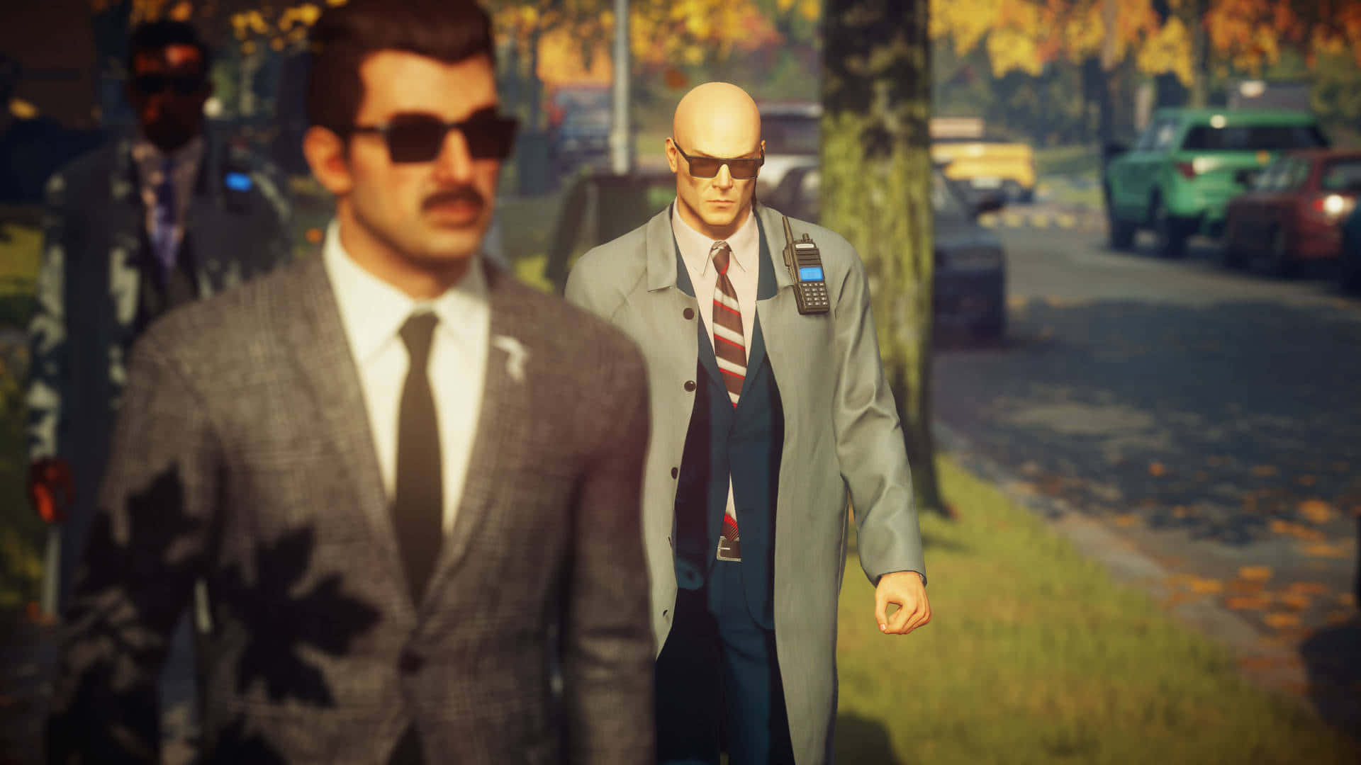 以高畫質探索《Hitman 2》的世界 壁紙 — 一個光頭男人穿著灰色乾濕褸和太陽眼鏡，走在兩旁種滿樹木的街道上，周圍有秋天的落葉飄落。