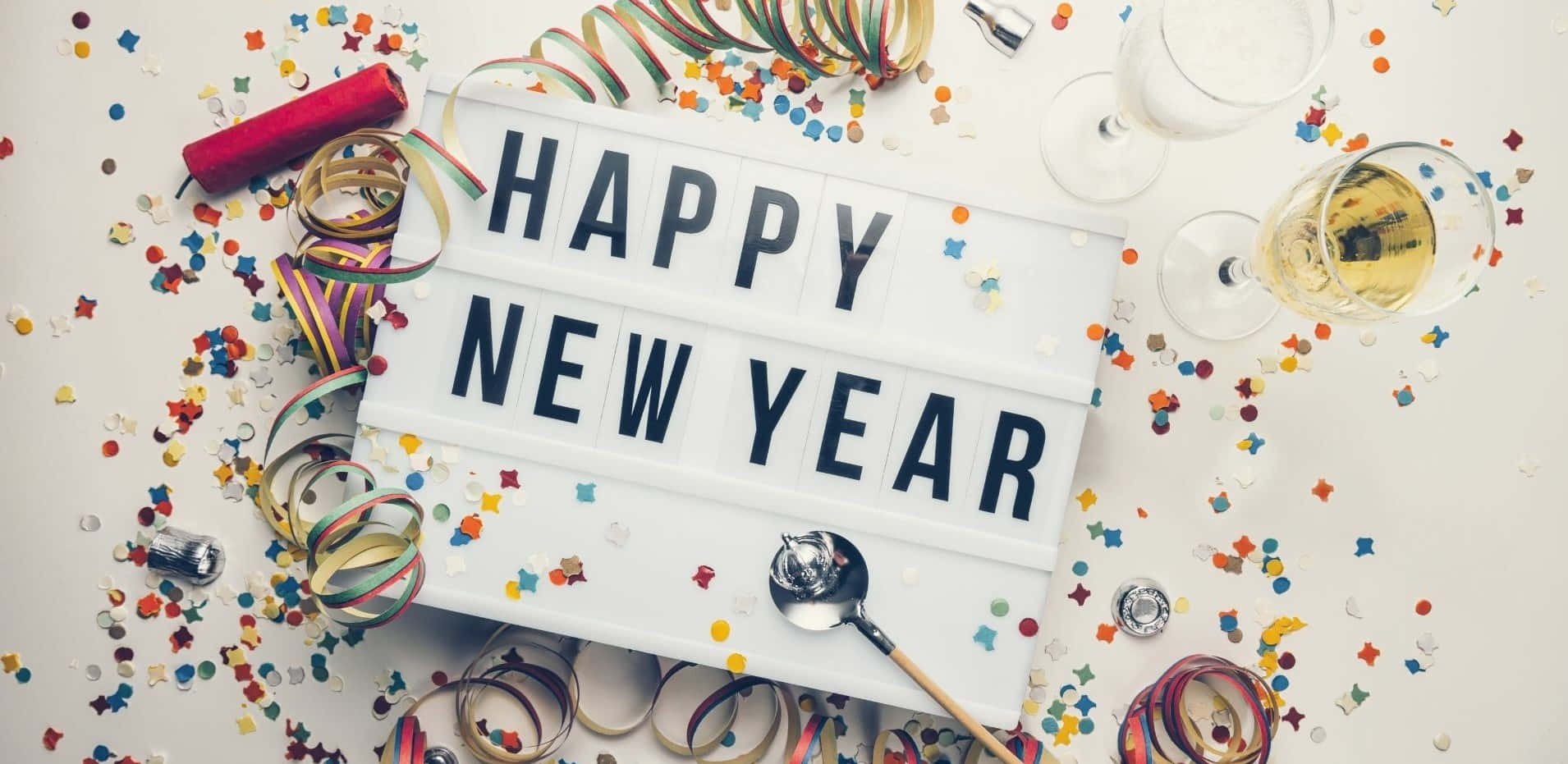 以鮮豔色彩慶祝新年！ 壁紙 — 一個節日白色燈箱，顯示「HAPPY NEW YEAR」，周圍環繞著五彩紙屑、絲帶、香檳杯和開瓶器，位於明亮的表面上。