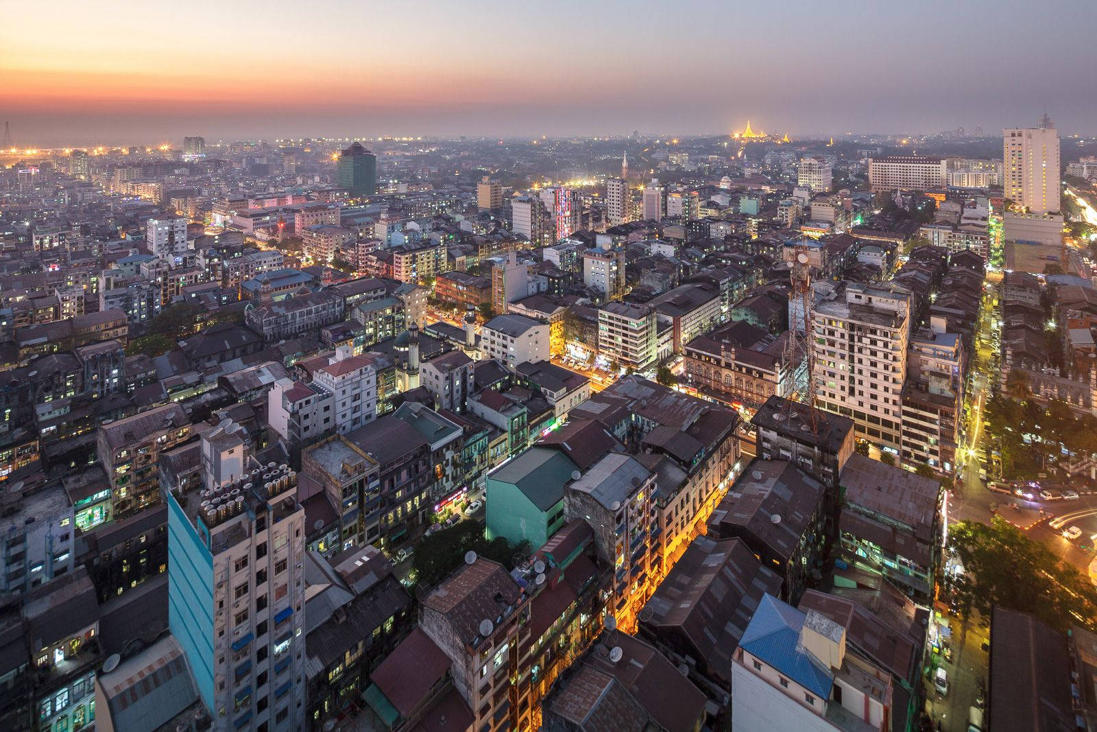 仰光夜景 壁紙 — 黃昏時分俯瞰一座廣闊的城市，溫暖的燈光從建築物和街道中閃耀，天空呈現柔和的橙色和紫色。