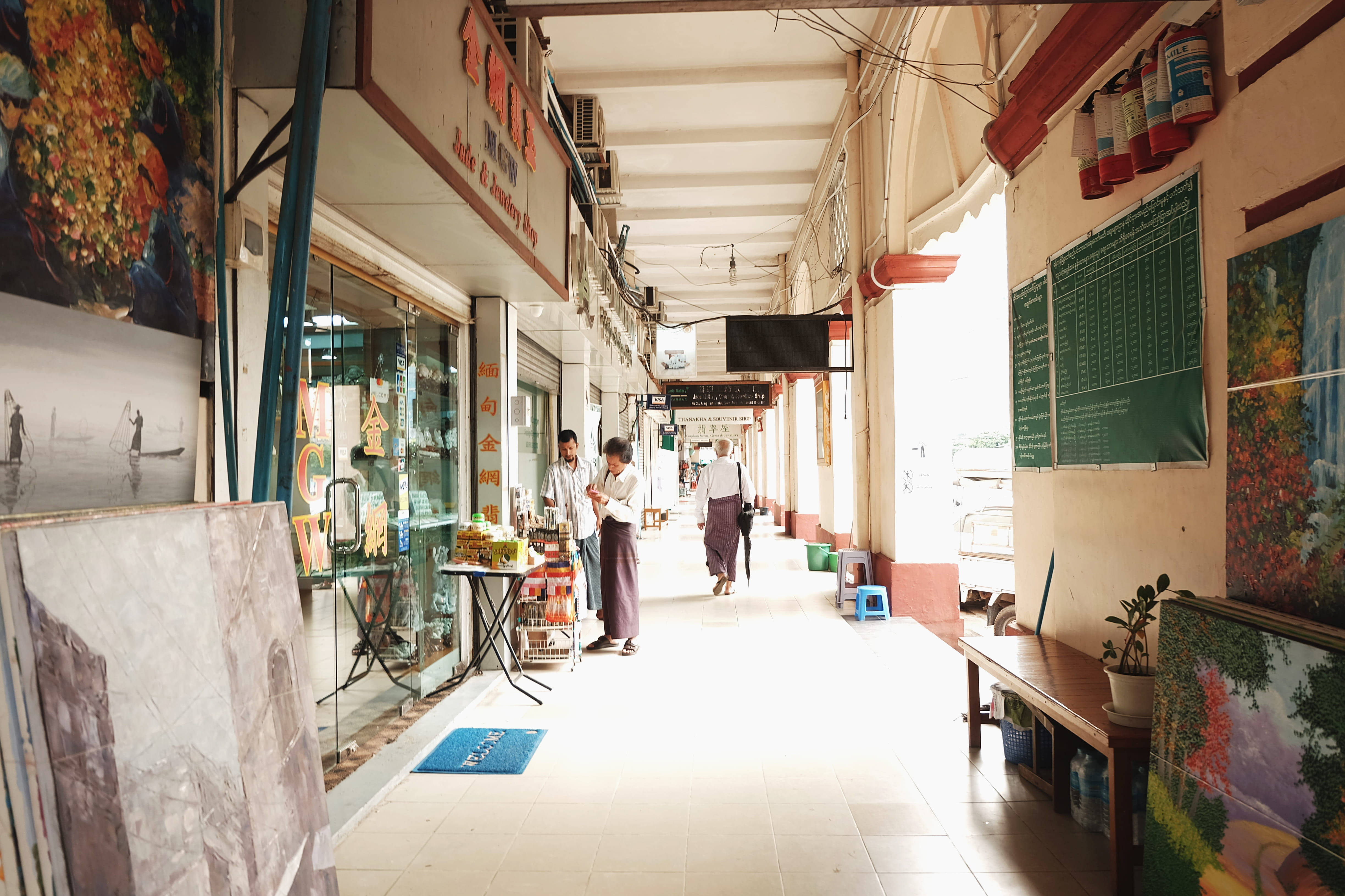 仰光市商業大廈遮蓋走廊 壁紙 — 一個明亮的帶頂棚小巷，在熙熙攘攘的市場中，有攤販和購物者在溫暖的陽光下。