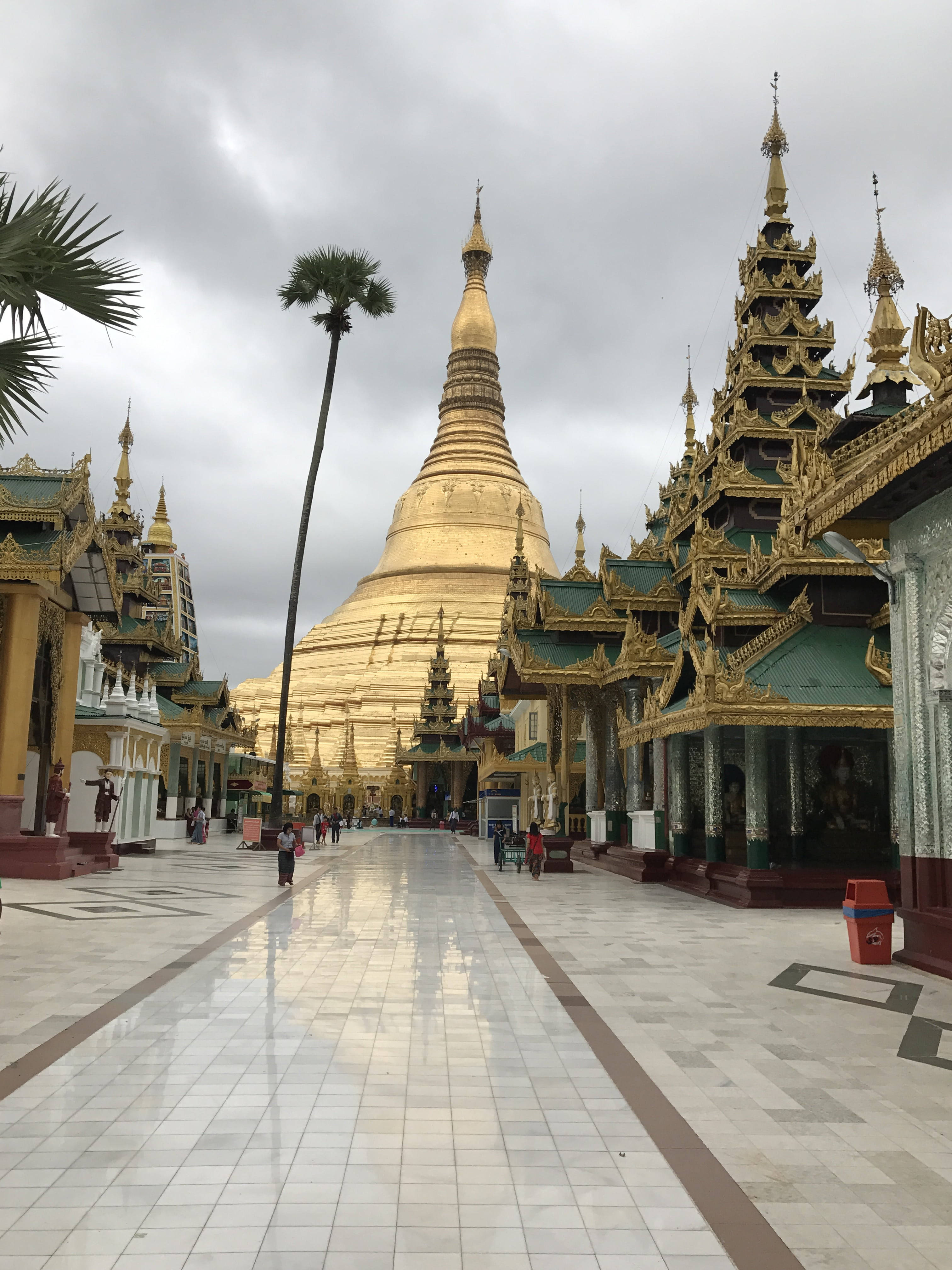 仰光錫塔戈恩寺廟區 壁紙 — 一座宏偉的金色寶塔在陰天聳立，兩旁是華麗的寺廟和棕櫚樹，濕漉漉的瓷磚步道延伸其間。