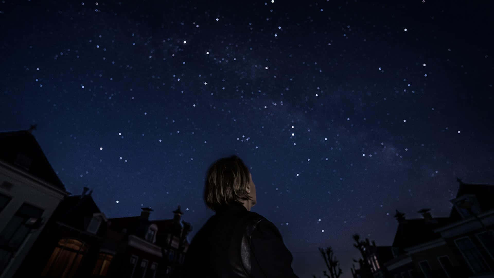 仰望星辰 壁紙 — 一個人抬頭仰望一排昏暗房屋上方、深邃藍色蒼穹下的繁星夜空。