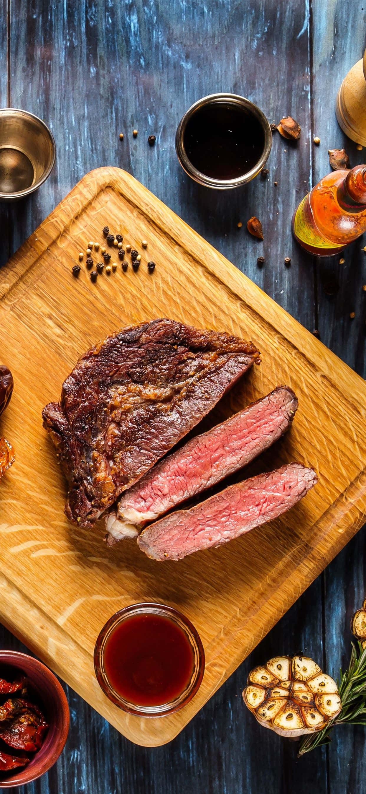 任何場合都適用的誘人美食 壁紙 — 完美煎烤的 medium-rare 牛排，切片放在木板上，配上大蒜、迷迭香和醬汁，置於鄉村風格的藍色桌子上。