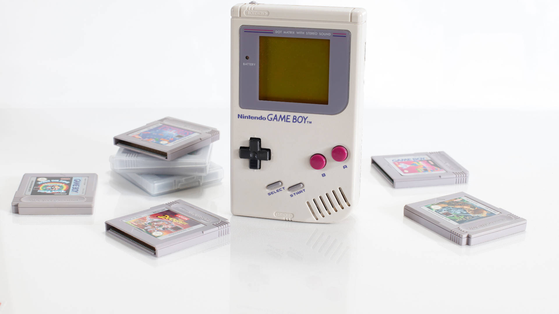 任天堂Game Boy與遊戲卡帶 壁紙 — 一部白色任天堂Game Boy配粉紅色按鈕，置中放在散落遊戲卡帶的反射白色表面上。