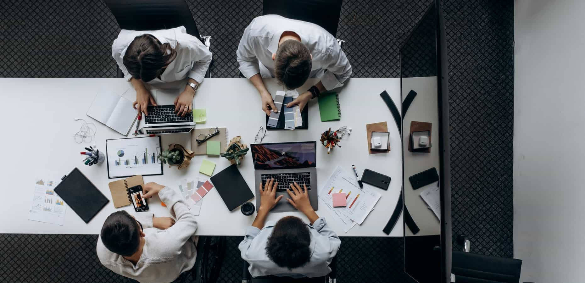 企業公司會議 壁紙 — 從上方俯瞰四人在現代辦公室的白桌上協作，桌上放有手提電腦、筆記本和設計材料。