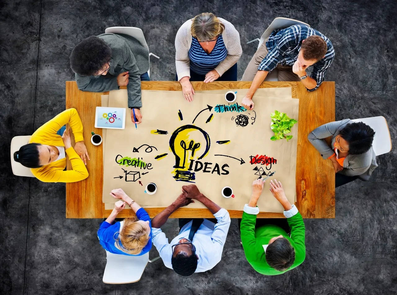 企業同事大腦風暴 壁紙 — Overhead view of diverse team brainstorming creative ideas around a wooden table with a large lightbulb sketch.