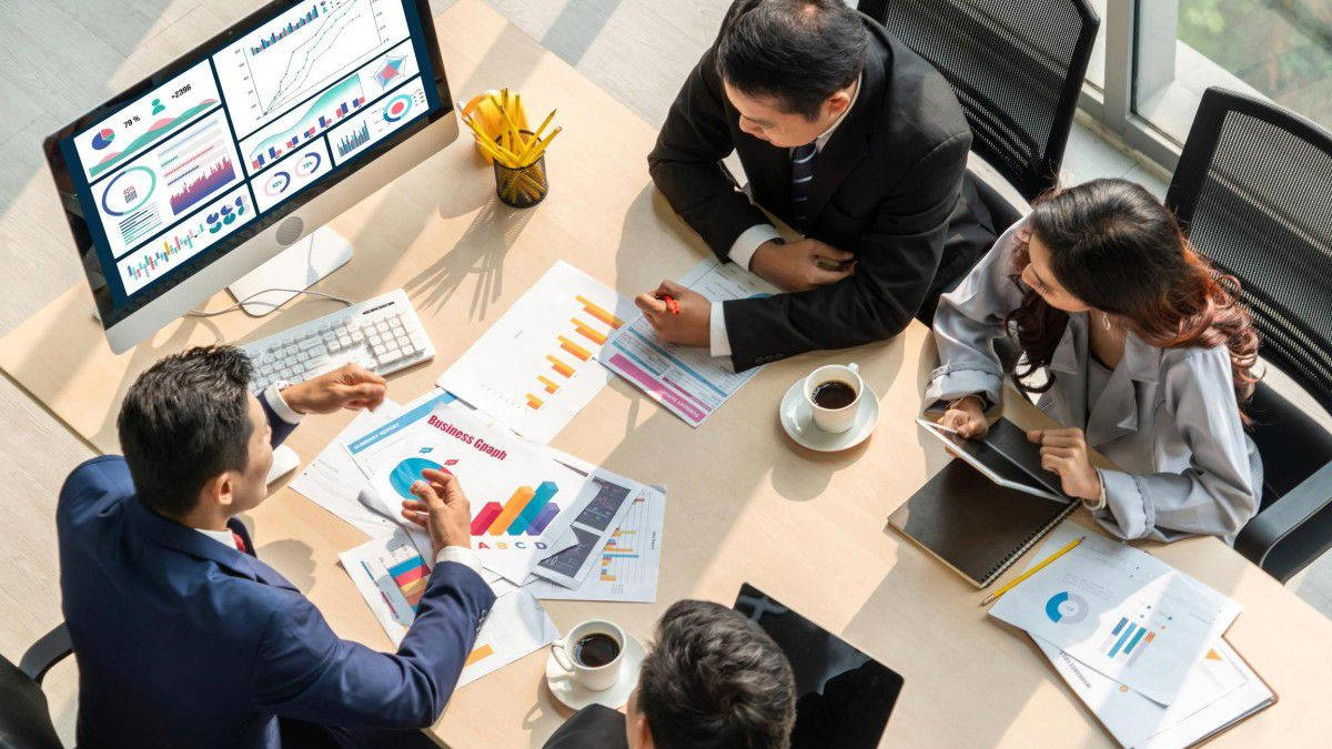 企業團隊進行戰略會議 壁紙 — Overhead view of four business professionals in suits collaborating around a conference table filled with colorful charts and data visualizations.