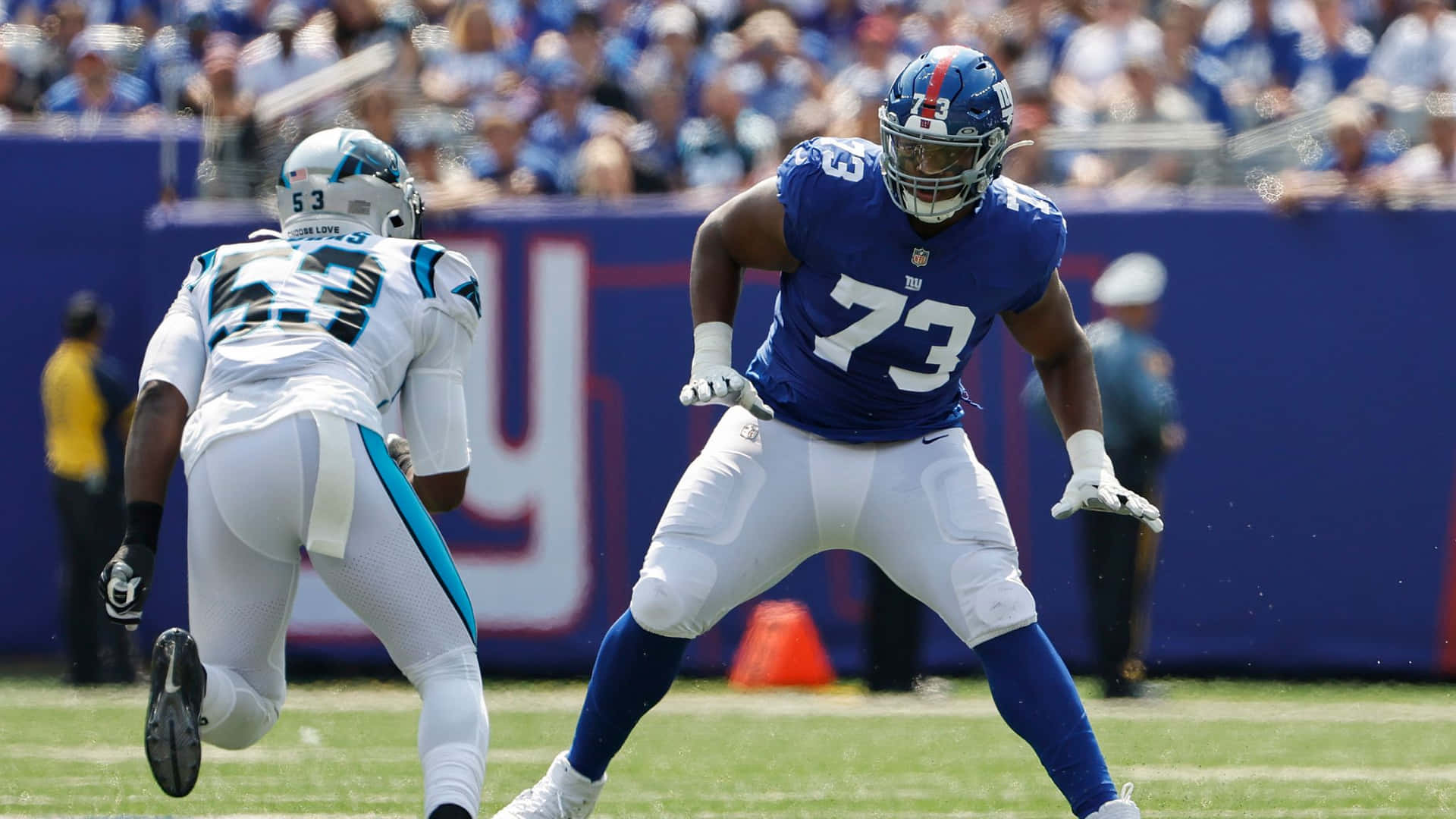 伊凡·尼爾進攻線衛 壁紙 — 兩名身穿藍白球衣的NFL球員在陽光照耀的草地上激烈競爭，背景有觀眾。