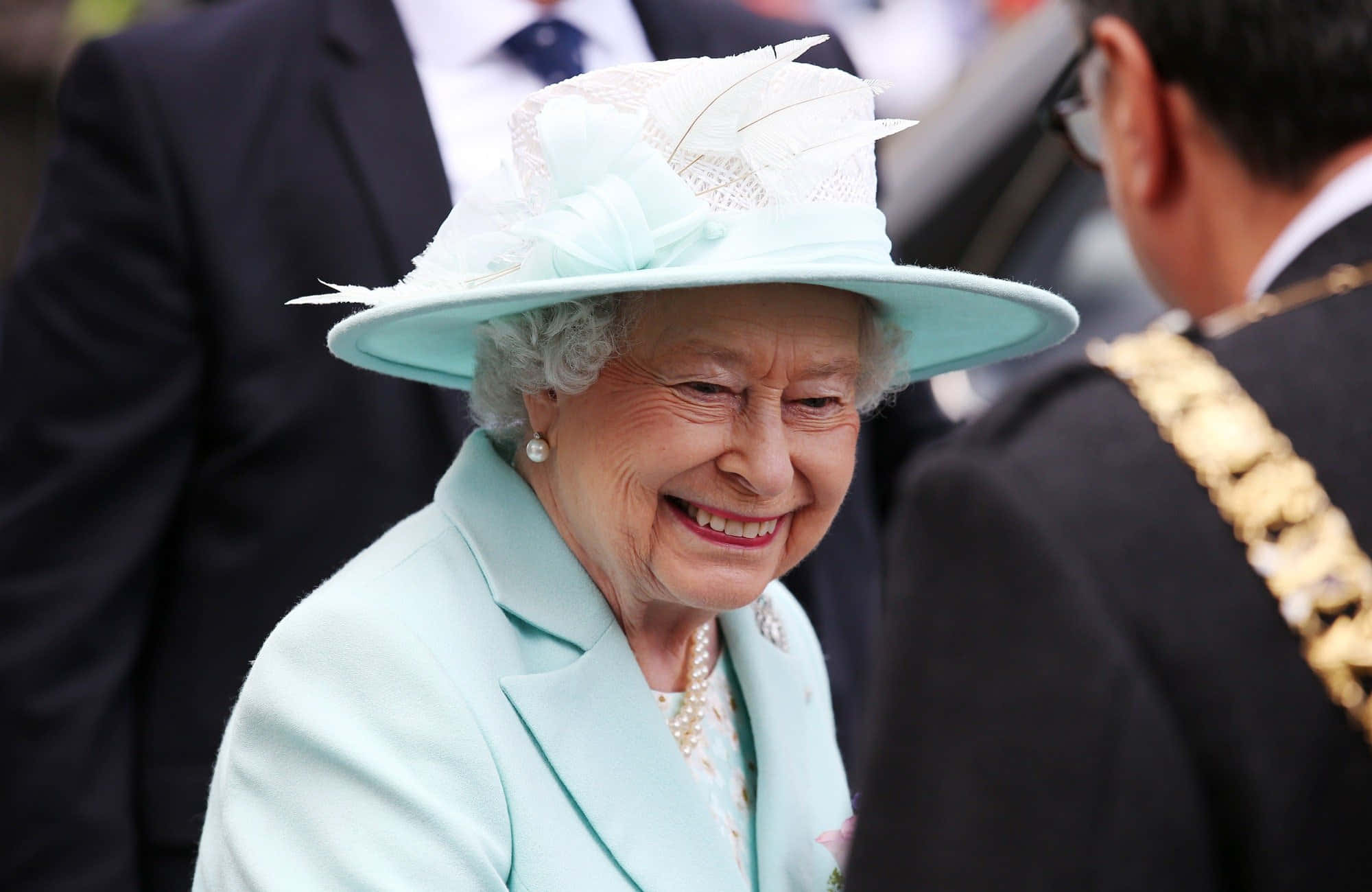 Queen Elizabeth 桌布 壁紙 — 伊利沙伯二世女王在公開活動中身穿淺藍色套裝和羽毛帽，溫暖地綻放笑容。